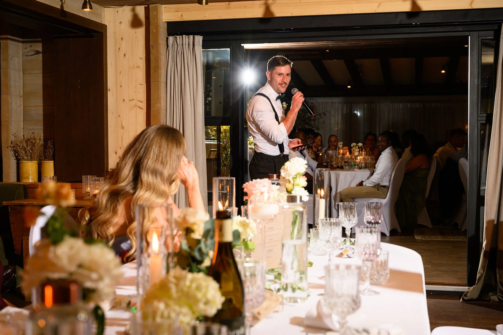 Un homme debout, portant une chemise blanche et un nœud papillon noir, parle dans un micro lors d'un événement, probablement un mariage, dans un restaurant ou une salle de réception, avec des invités assis à des tables décorées avec des fleurs et des