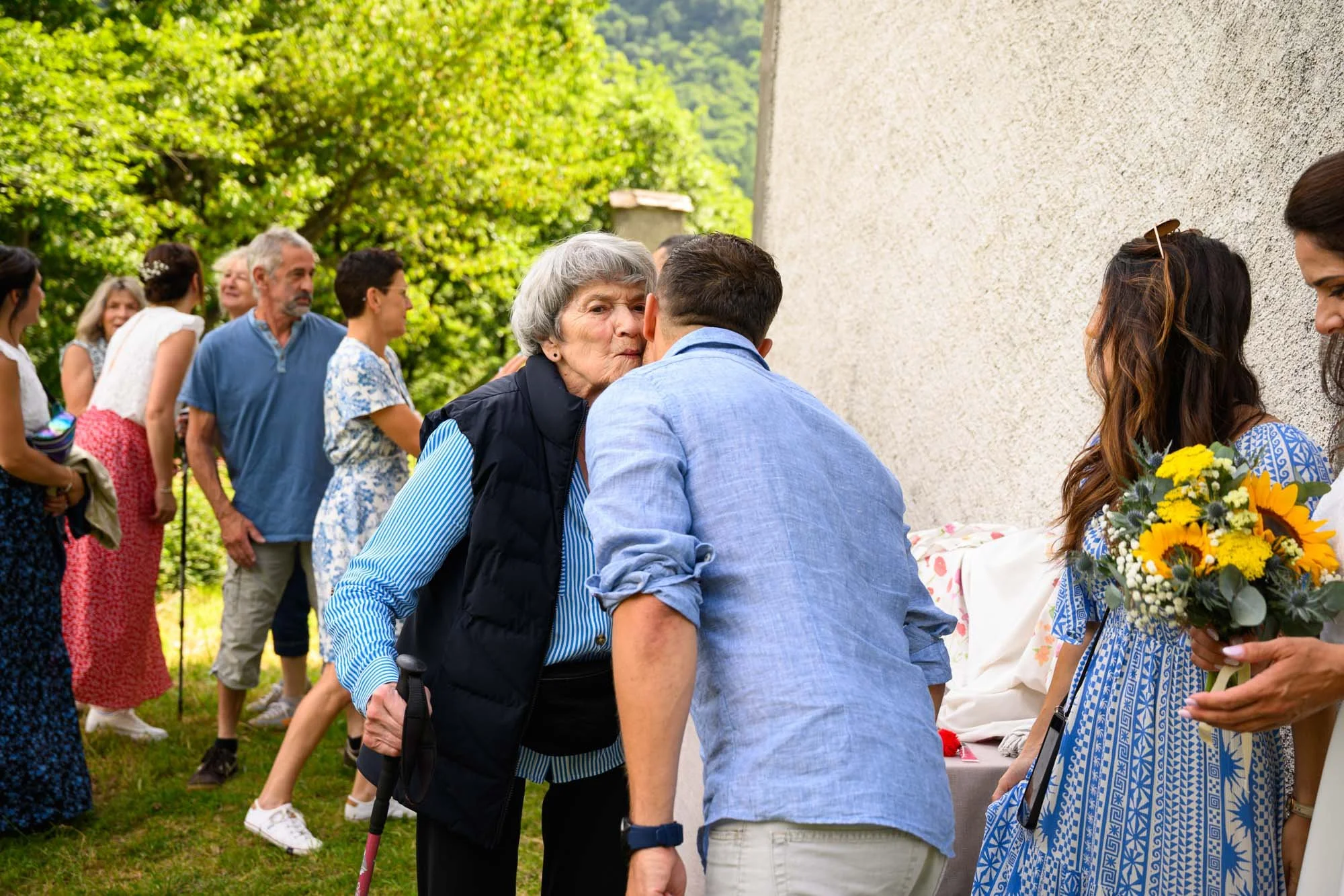 Un groupe de personnes à une fête en plein air, avec une femme âgée embrassant un homme, et d'autres personnes en arrière-plan, certains tenant des fleurs.