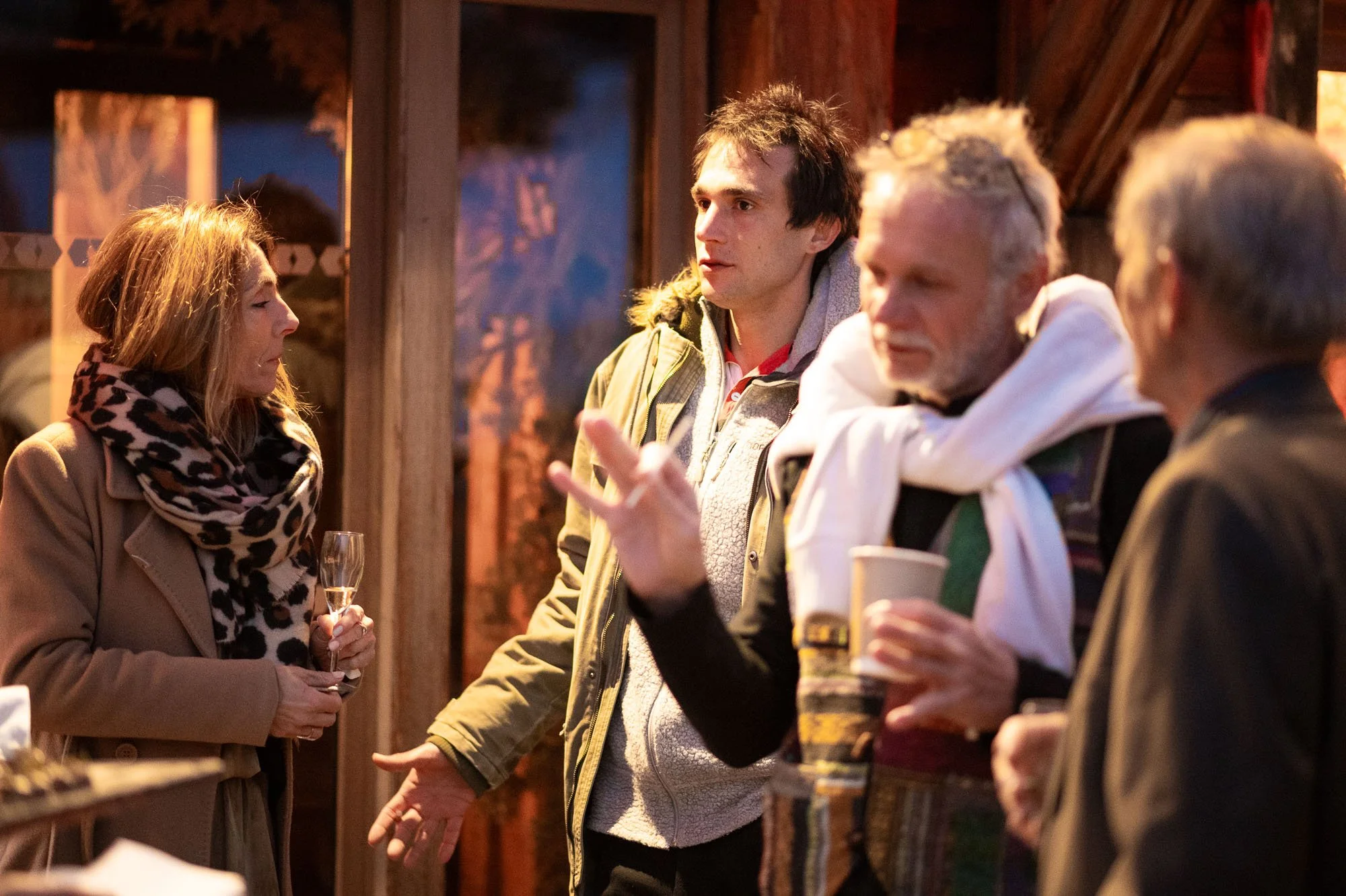 Groupe de quatre personnes discutant lors d'une soirée, une femme avec un foulard à motifs, un homme avec une veste beige, un homme avec une écharpe et une tasse, et un homme avec des cheveux gris, dans un cadre chaleureux.