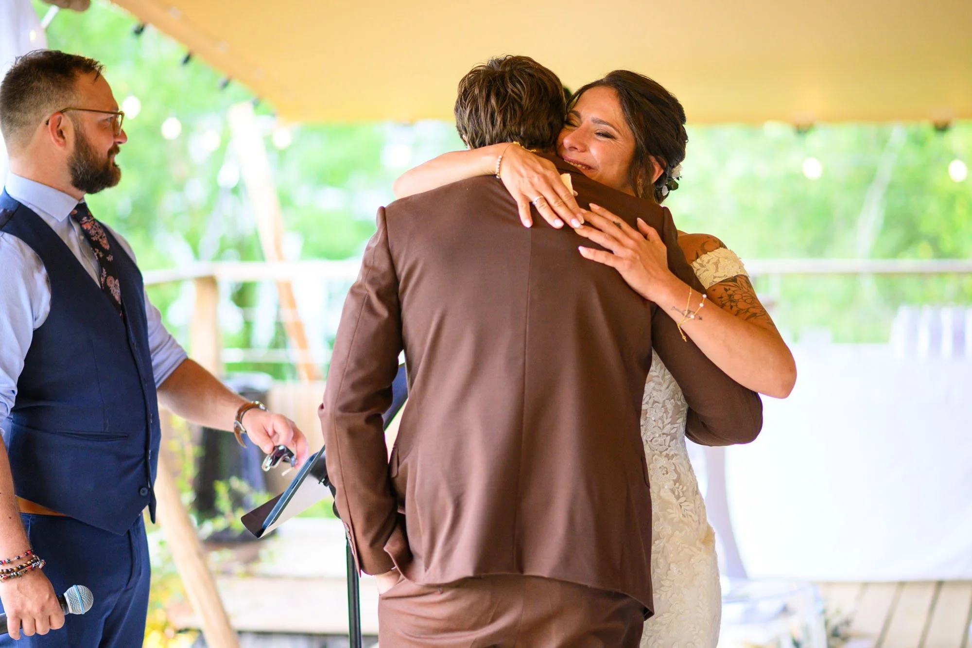 Deux personnes s'embrassent lors d'une cérémonie de mariage en plein air, une femme en robe de mariée souriante et un homme en costume marron, un homme à gauche tient un micro.