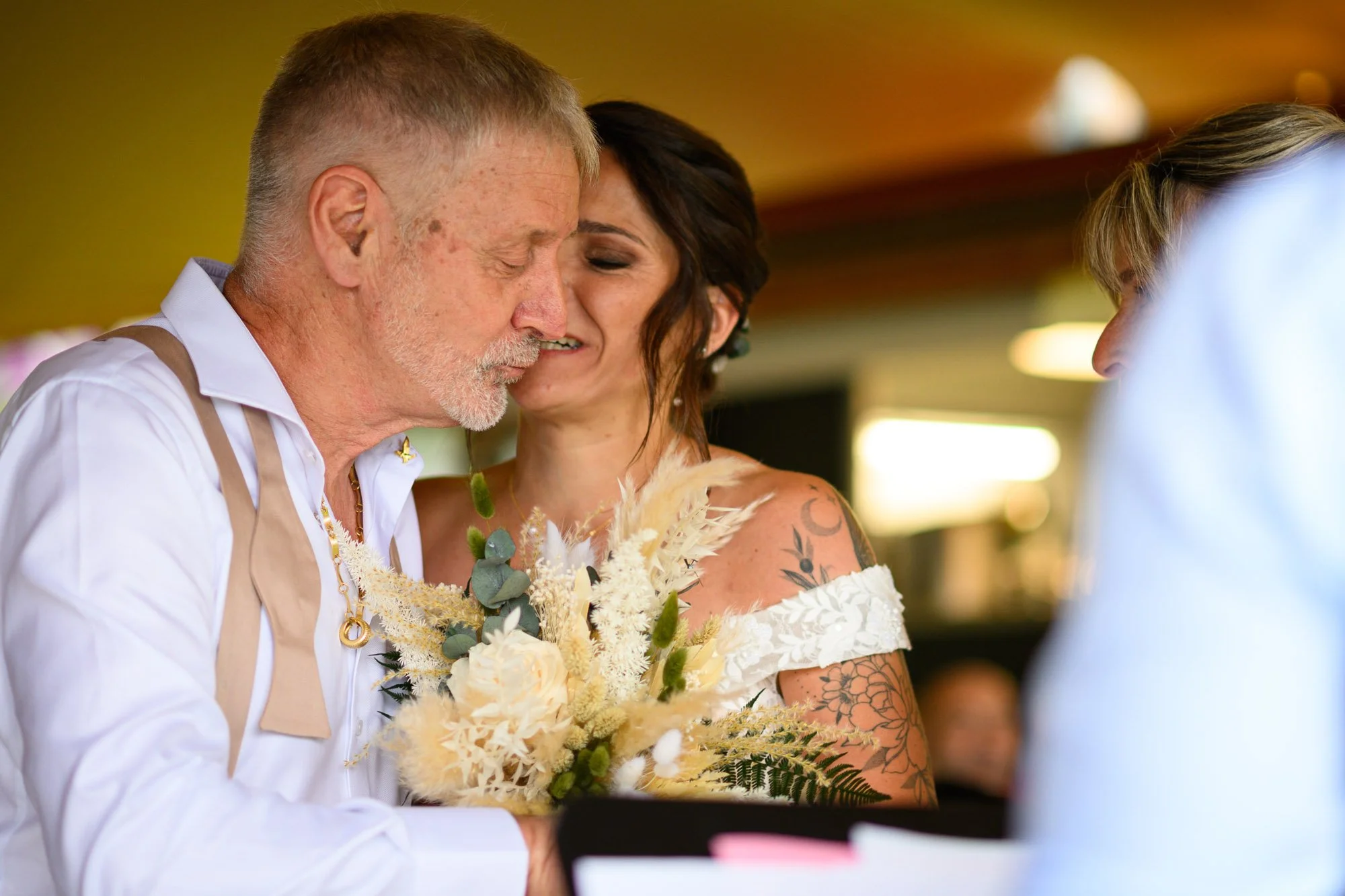 Un couple échange des vœux de mariage, avec le marié plus âgé et la mariée portant un bouquet de fleurs, lors d'une cérémonie en intérieur.
