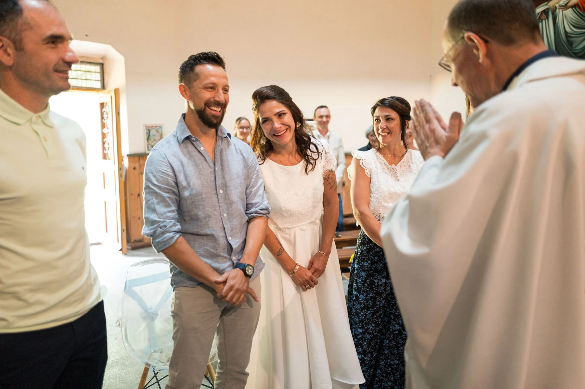 Un groupe de personnes souriantes lors d'une cérémonie religieuse, probablement un mariage, dans une salle avec un prêtre ou un officiant en robe blanche.