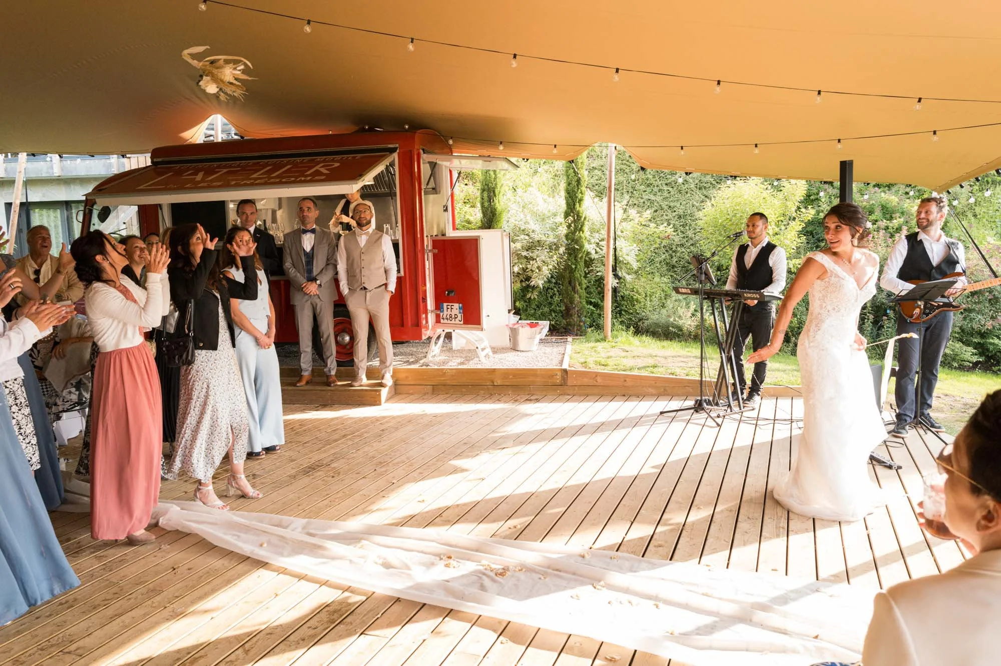 Une mariée danser avec un groupe de gens lors d'un mariage en extérieur, avec un groupe de musiciens en arrière-plan, sous une grande tente beige, entourée de arbres verts.