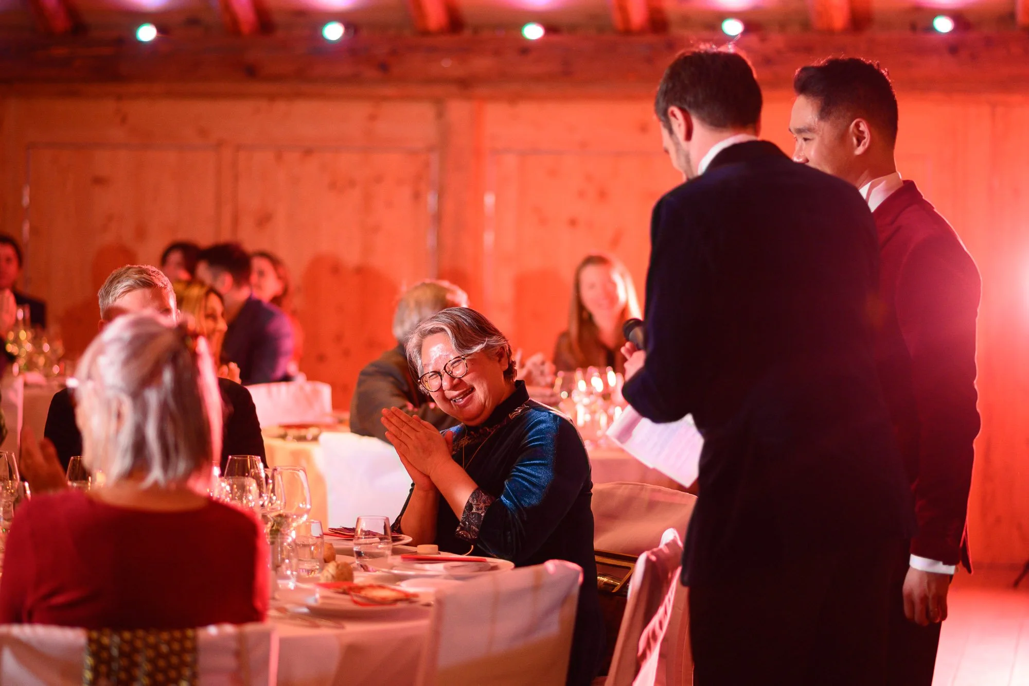 Une femme souriante et ravie écoute un discours lors d'une cérémonie ou d'un dîner, avec un groupe d'invités assis autour d'une table élégamment dressée dans un décor en bois, sous un éclairage chaud.
