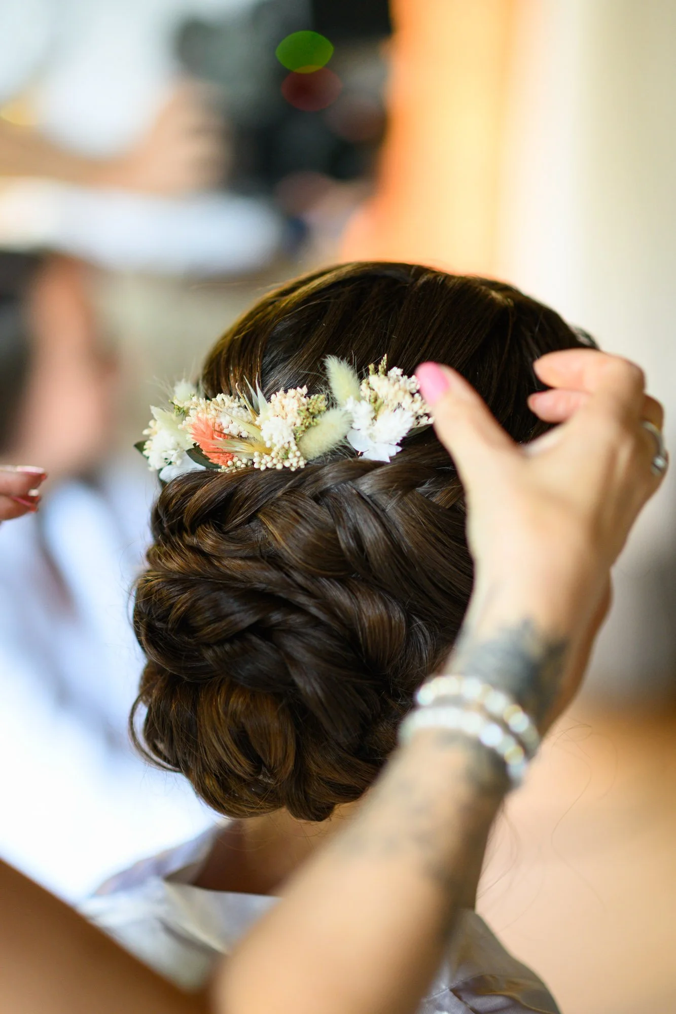 Femme avec une coiffure tressée portant une couronne de fleurs, en train de se faire préparer pour un événement. Le focus est sur la coiffure et la couronne, avec une personne aidant à ajuster. Décor flou en arrière-plan.