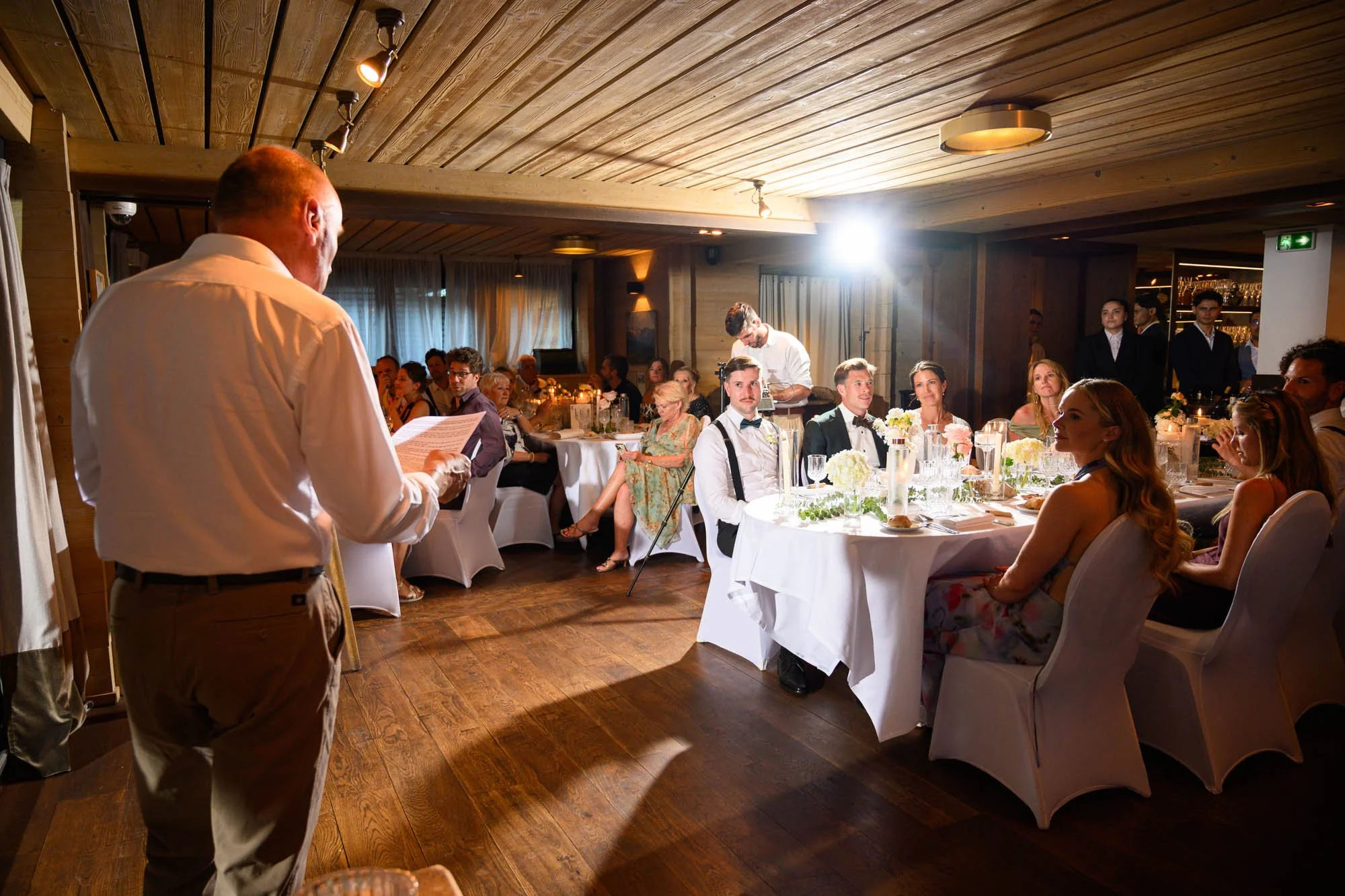 Discours lors d'un mariage dans une salle élégante avec des invités assis autour de tables décorées de fleurs et de vaisselle raffinée.