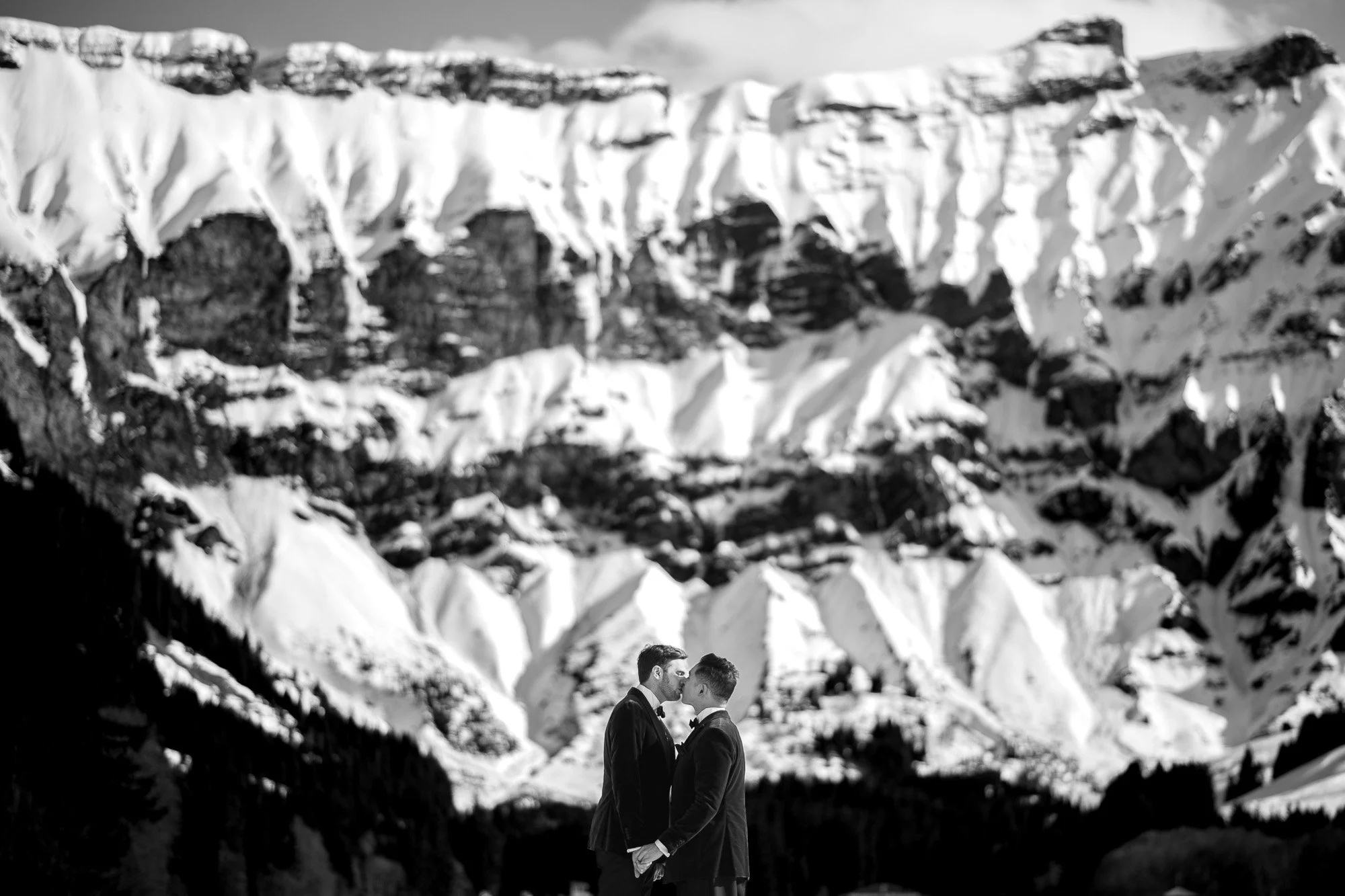 Deux hommes en costume se tiennent face à face en se tenant par la main, dans un paysage montagneux enneigé avec des falaises abruptes en arrière-plan.