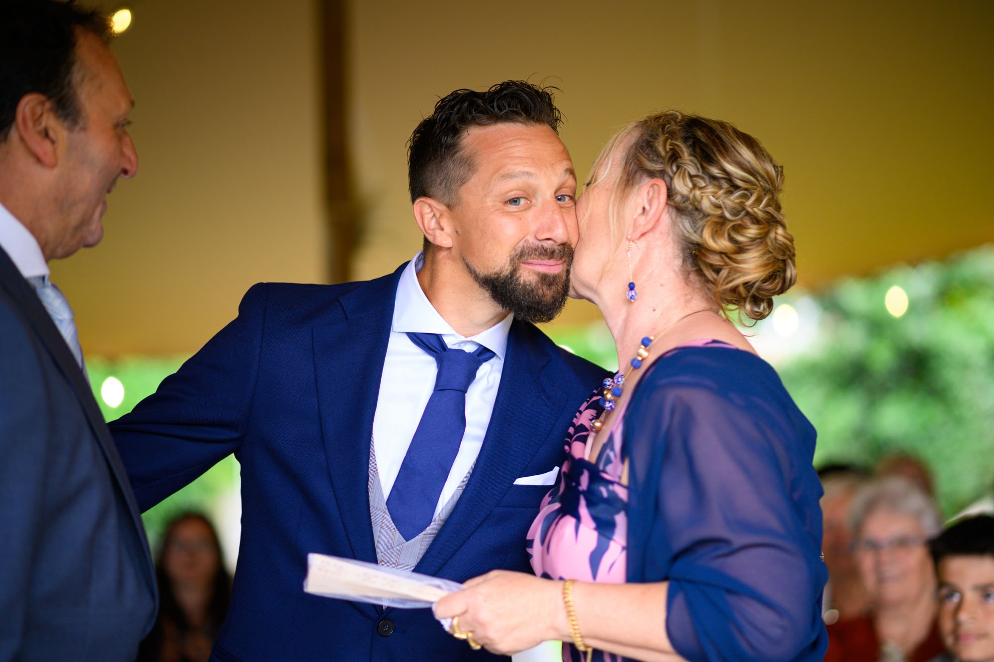 Une femme donne un baiser sur la joue à un homme lors d'une cérémonie, probablement un mariage, avec d'autres personnes en arrière-plan.