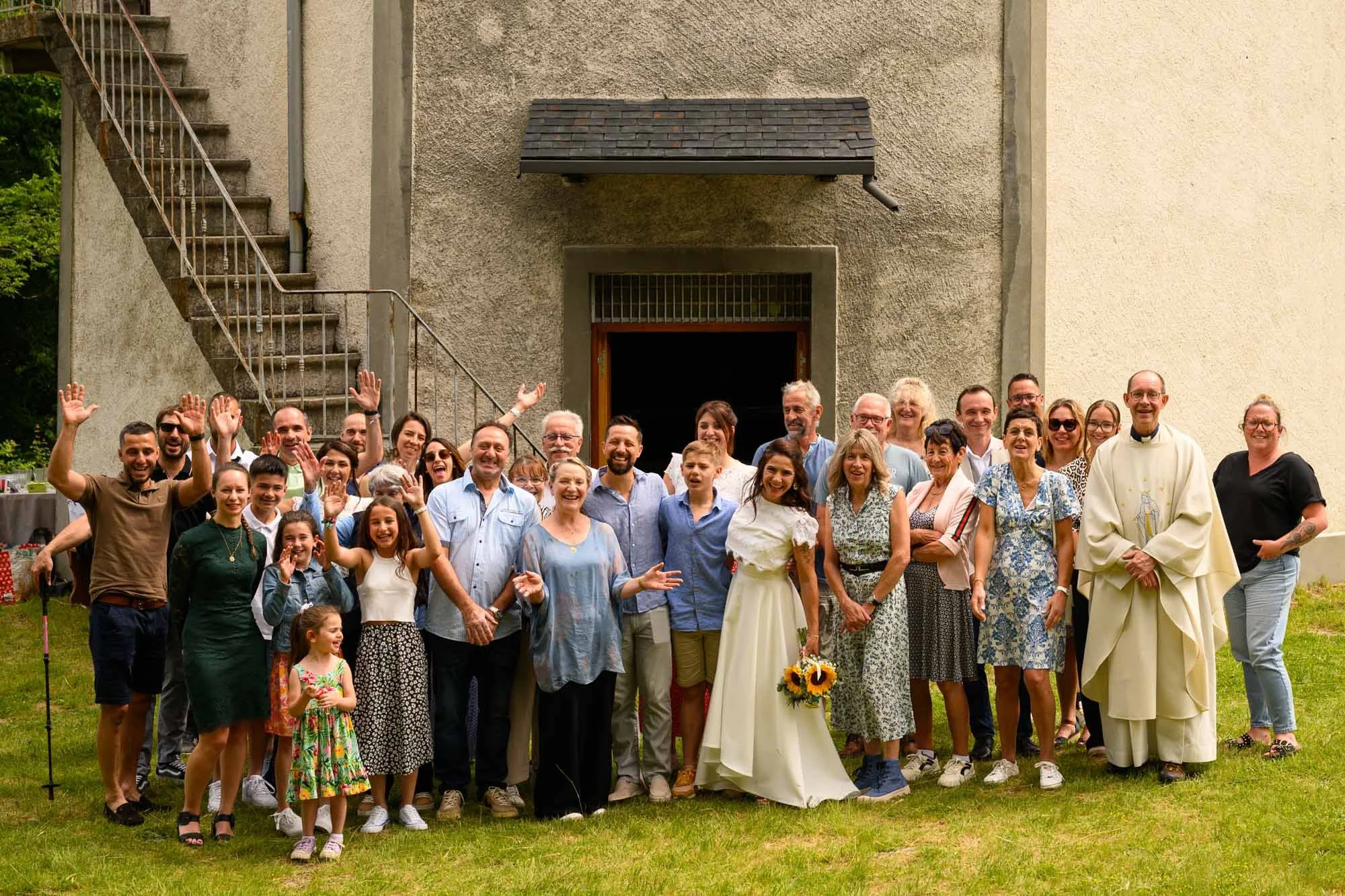 Groupe de personnes souriantes réunies à l'extérieur d'une église en pierre, célébrant un événement, avec un prêtre en robe blanche à droite.