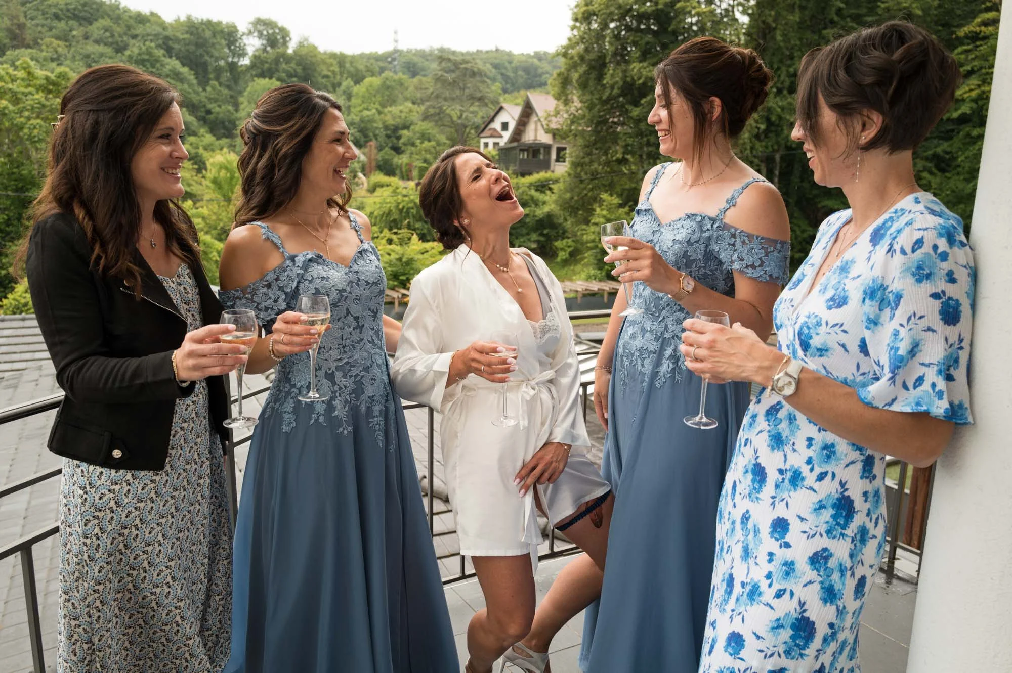Groupe de six femmes habillées élégamment, souriantes, en train de discuter et de boire du champagne sur une terrasse avec un fond de verdure.