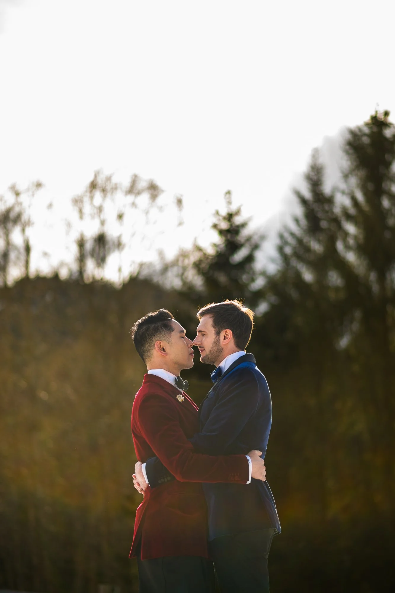 Deux hommes habillés en costumes élégants, se tenant proches avec leur front presque touchant, dans un cadre extérieur avec des arbres en arrière-plan, sous une lumière naturelle douce.