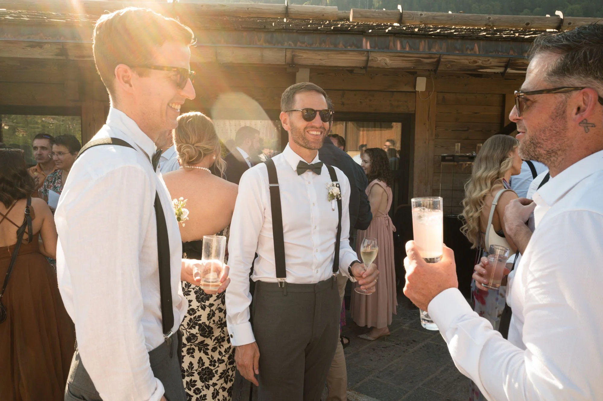 Groupe d'hommes en tenue de mariage levant leurs verres lors d'une réception en plein air, avec une ambiance chaleureuse et soleil couchant.
