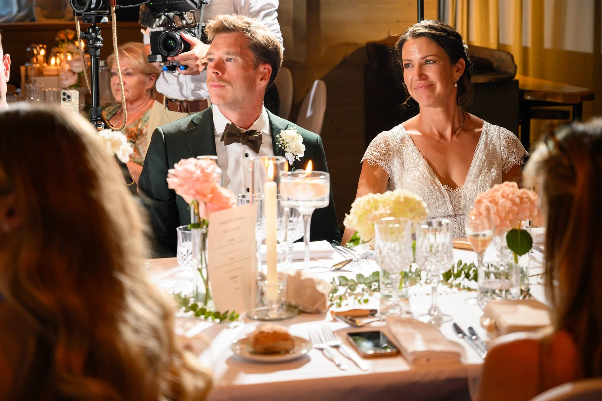 Un couple assis à une table lors d'un mariage, la femme souriante et l'homme en smoking avec un nœud papillon, entourés de convives et de décorations florales.