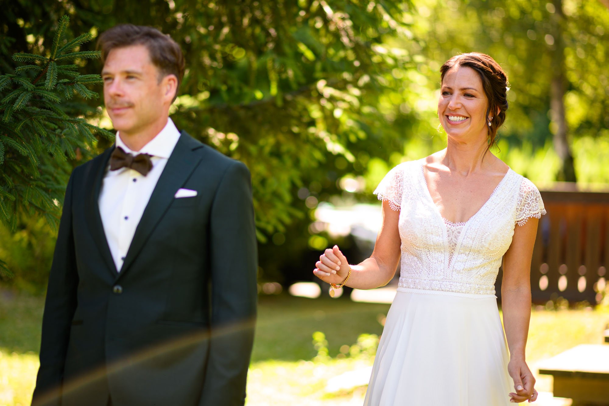 Un couple lors d'un mariage en plein air, l'homme en costume noir avec une chemise blanche et un nœud papillon, la femme en robe blanche avec des détails en dentelle, souriant dans un jardin ensoleillé avec des arbres en arrière-plan.