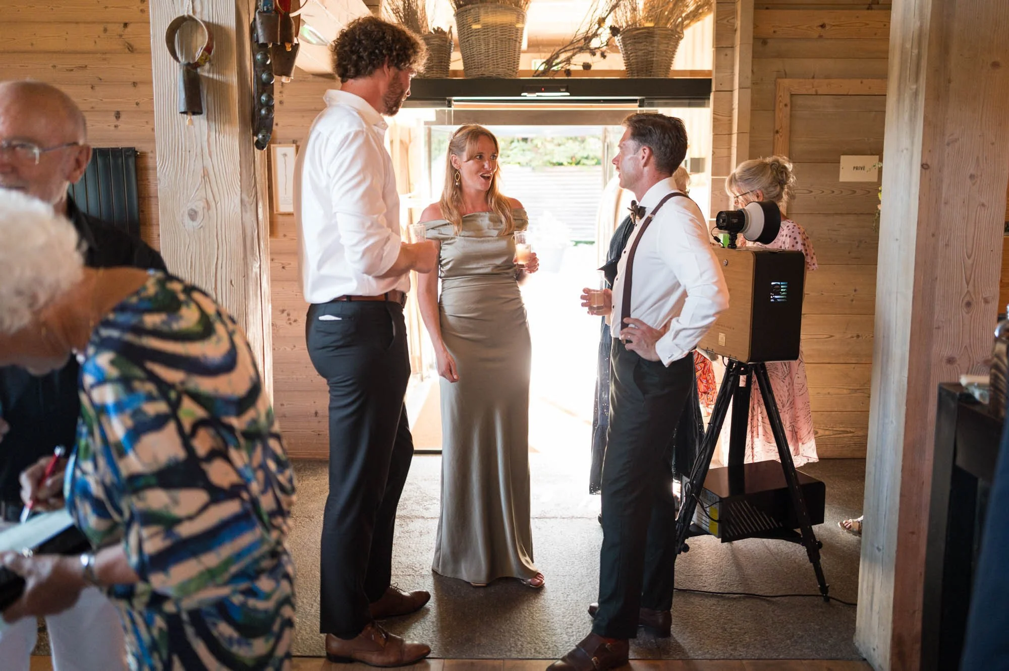 Groupe de personnes à une réception ou un événement social, discutaillant, avec décoration en bois et équipement audio visible.