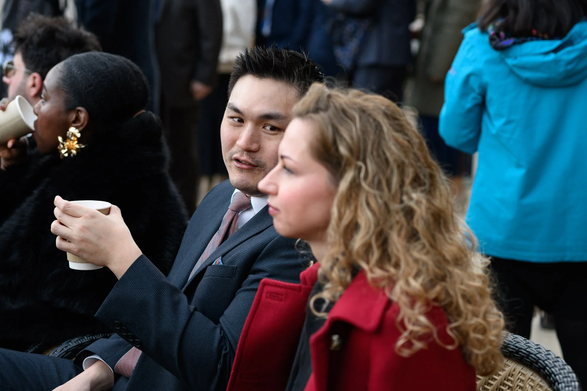 Deux personnes assises lors d'un événement ou d'une réunion, tenant une tasse en papier, avec d'autres personnes en arrière-plan.