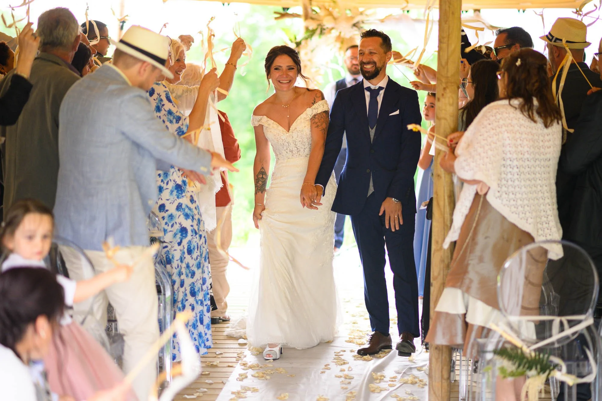 Un couple de mariés heureux marchant main dans la main au milieu d'une cérémonie de mariage en plein air, entouré d'amis et famille qui célébrent, sous une tente décorée de rubans et de fleurs.