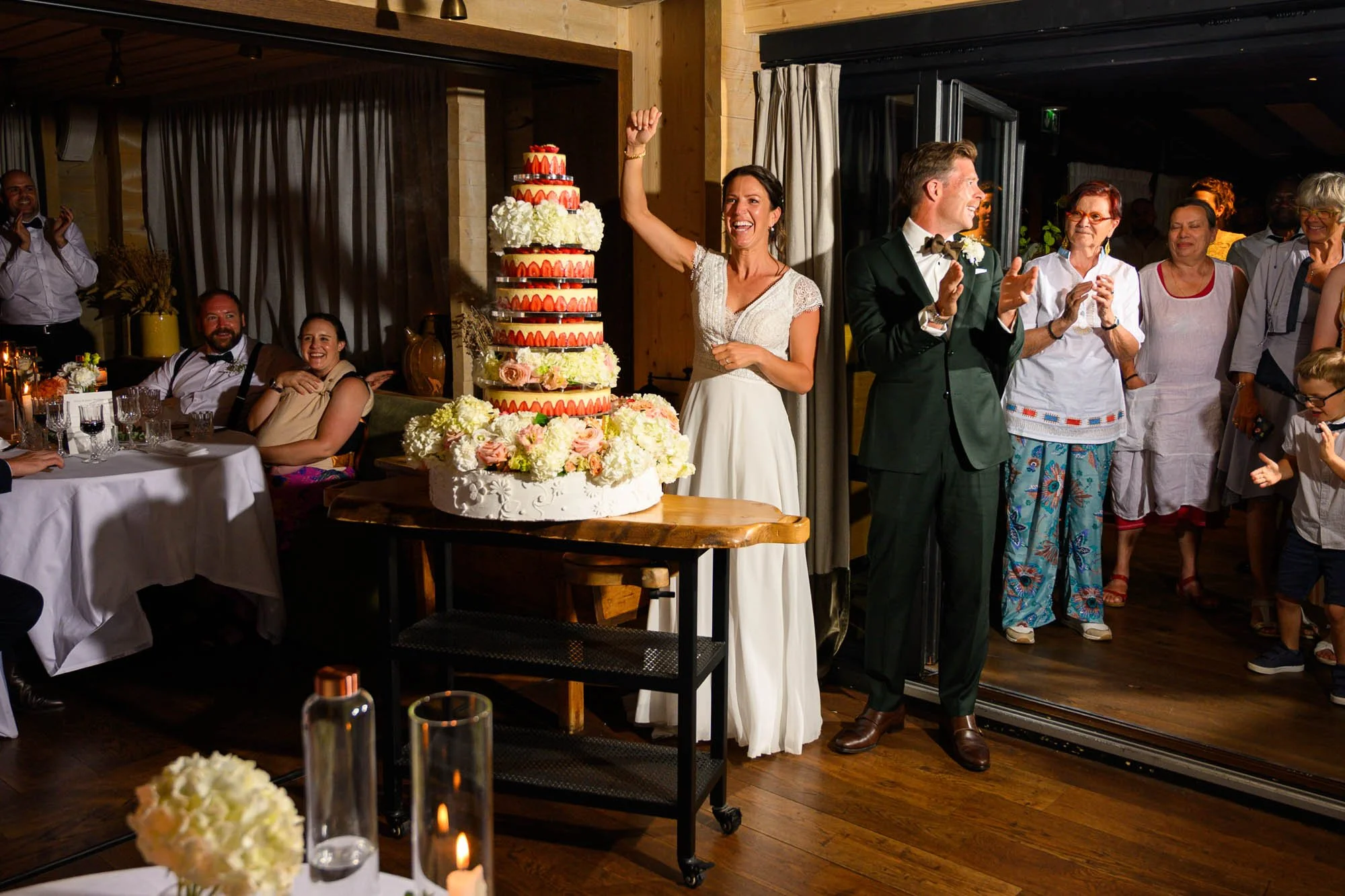 Mariée souriante avec bras levé célébrant à côté d'un grand gâteau blanc décoré de fleurs et de fraises, avec des invités applaudissant et souriant dans une ambiance chaleureuse et festive.