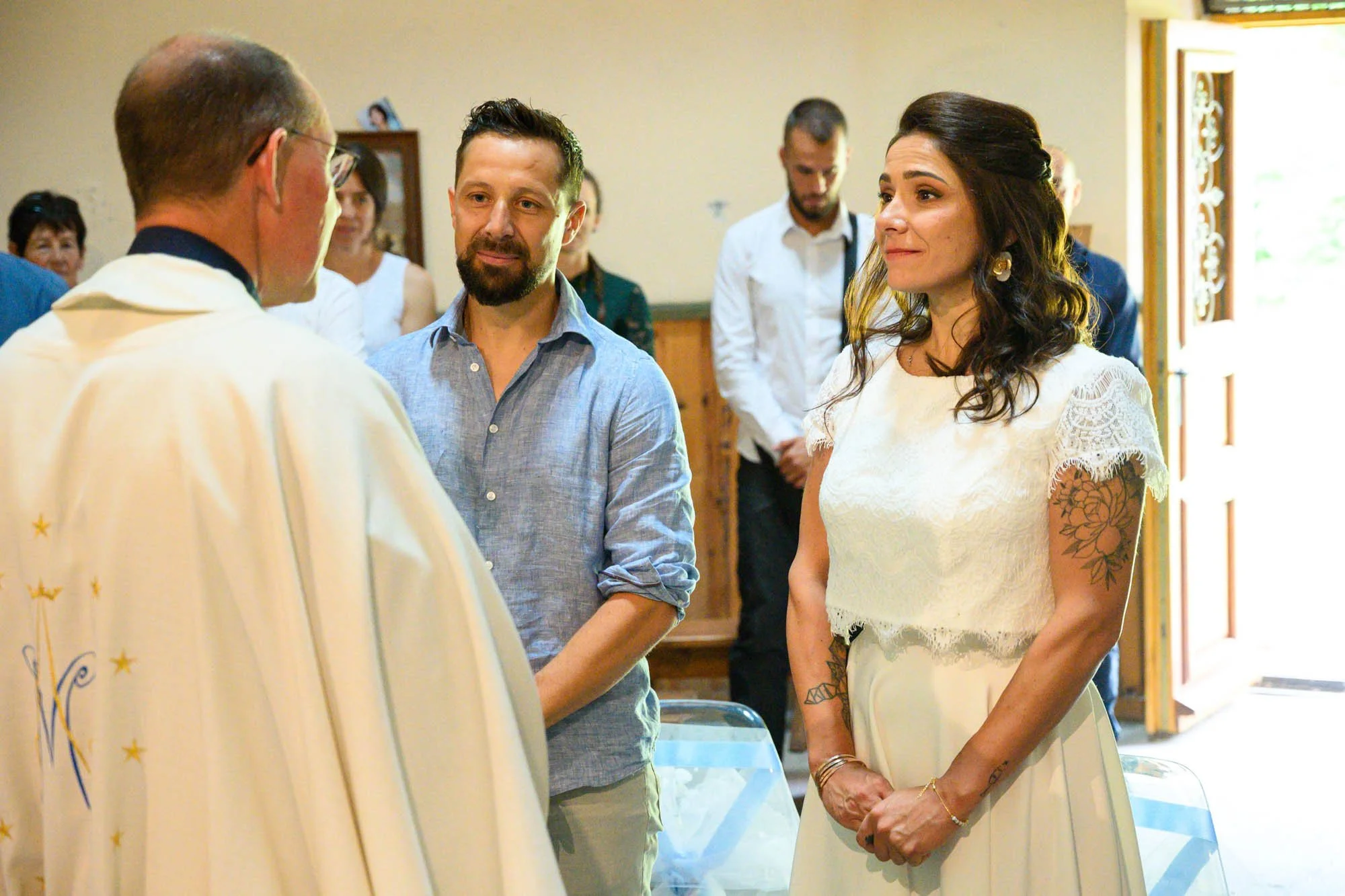 Une cérémonie religieuse où une femme en robe blanche avec tatouages et un homme en chemise bleue regardent un prêtre lors d'une cérémonie, avec d'autres personnes en arrière-plan.