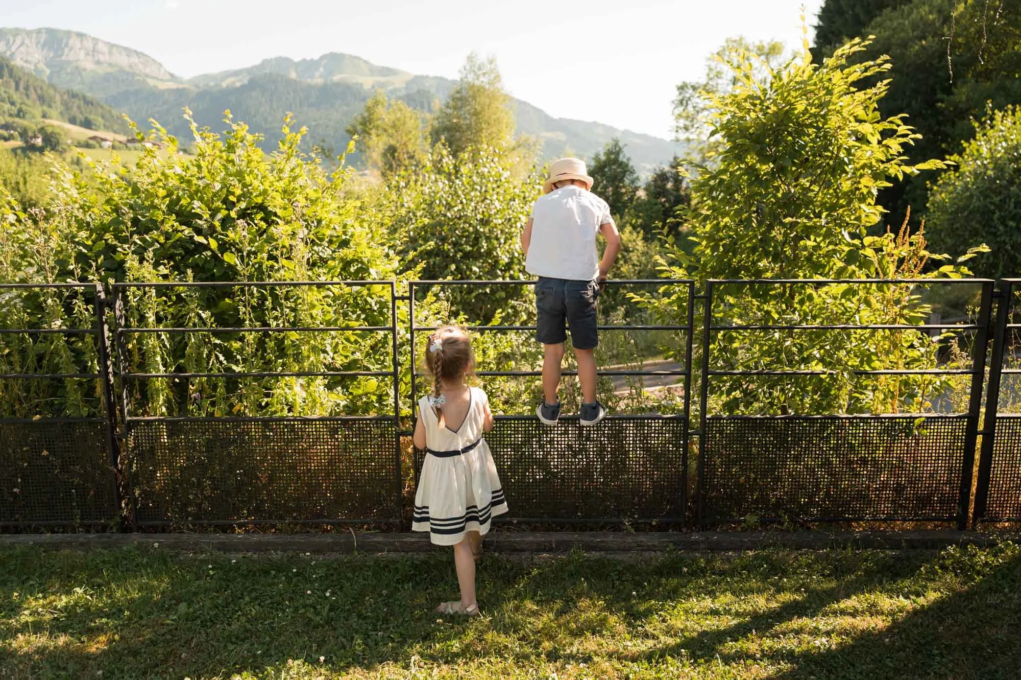 Deux enfants regardent à travers une clôture en métal vers un jardin avec des arbres et des montagnes en arrière-plan, soleil brillant, ambiance estivale.