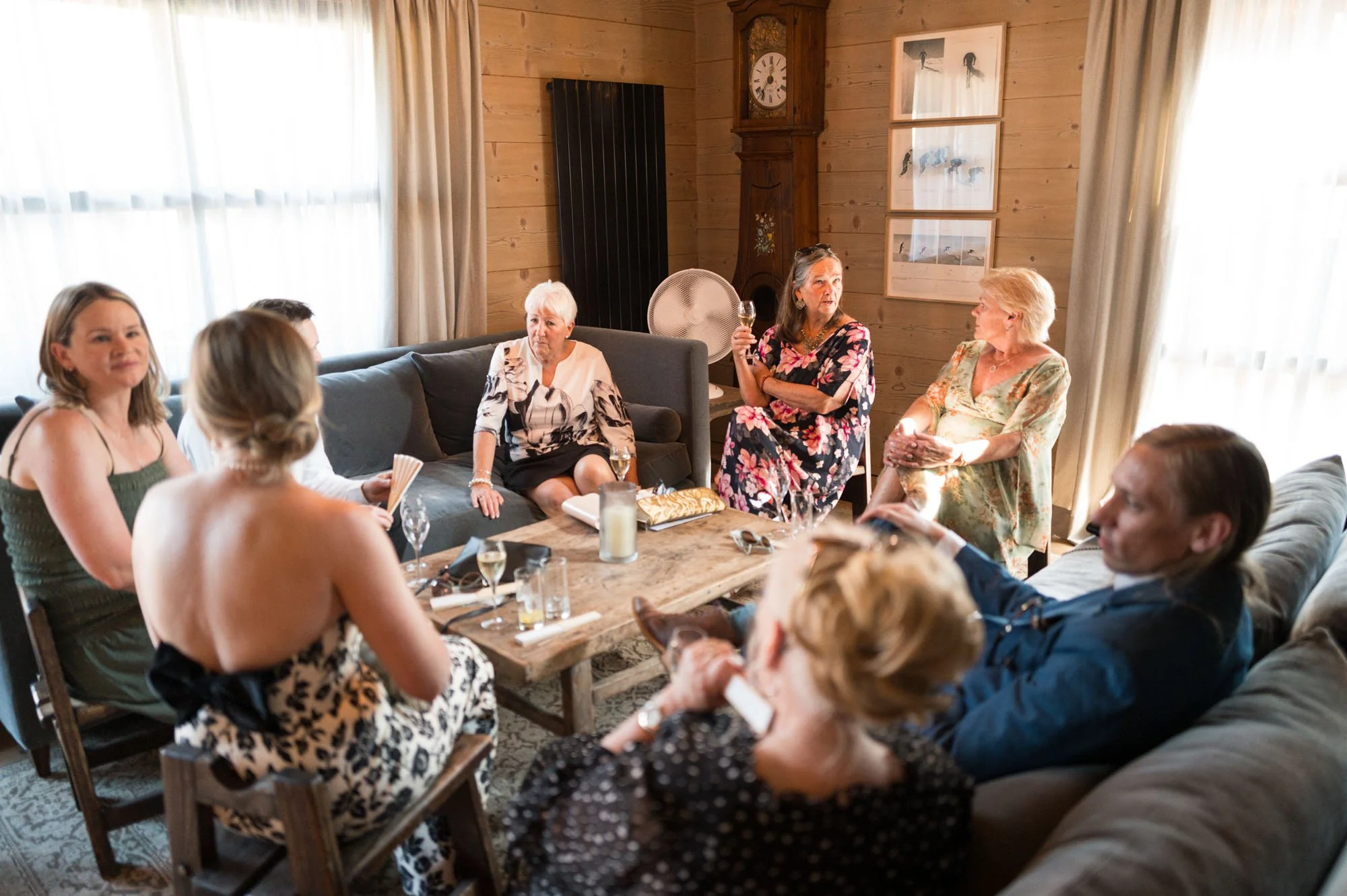 Groupe de personnes féminines et masculines assises dans un salon en bois, discutant lors d'une réunion ou d'une fête, quelques-unes tenant des verres et une personne au centre parlant.