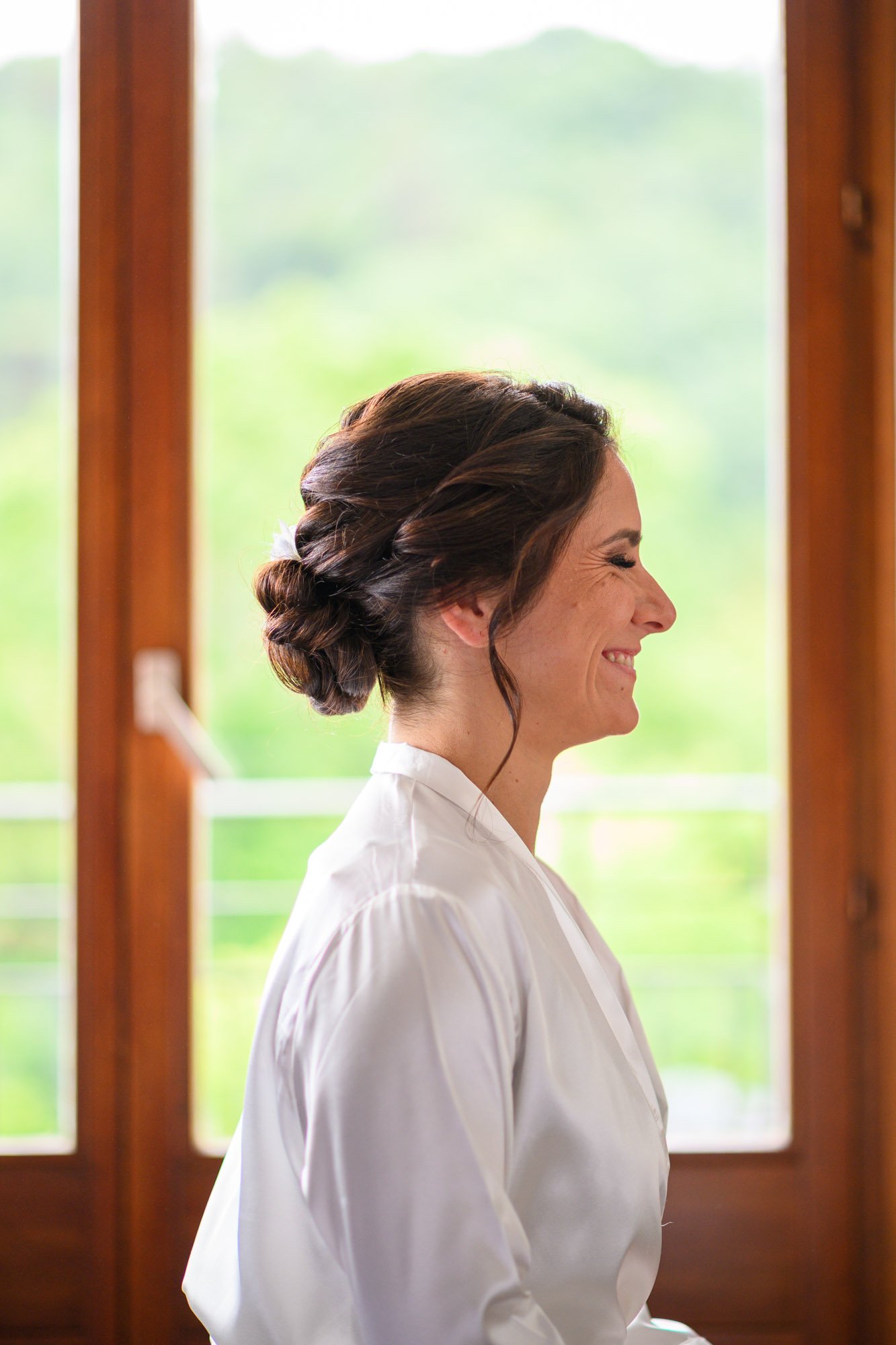 Une femme souriante en profil, avec ses cheveux brun tressés, portant une robe blanche, devant une fenêtre en bois avec un paysage vert flou en arrière-plan.
