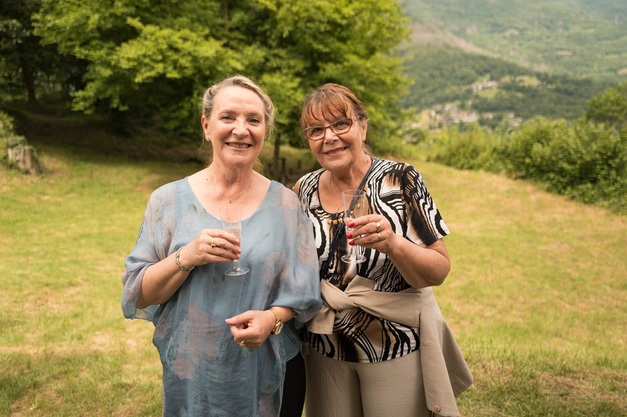 Deux femmes souriantes tenant des verres à champagne dans un champ verdoyant, avec des arbres et des montagnes en arrière-plan.