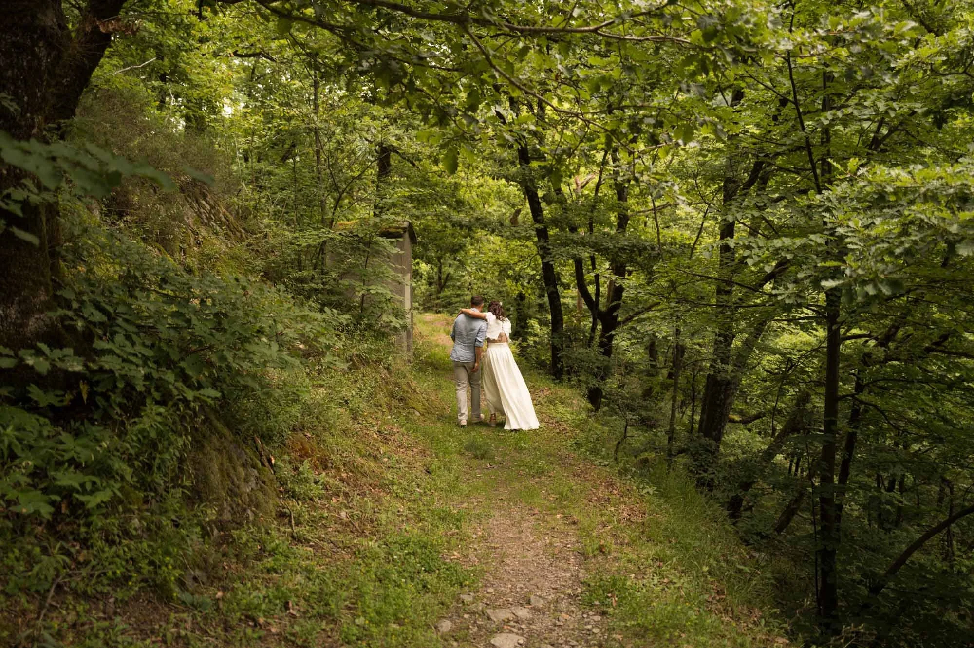 Un couple de mariés marche main dans la main dans une forêt verdoyante, vue de derrière.