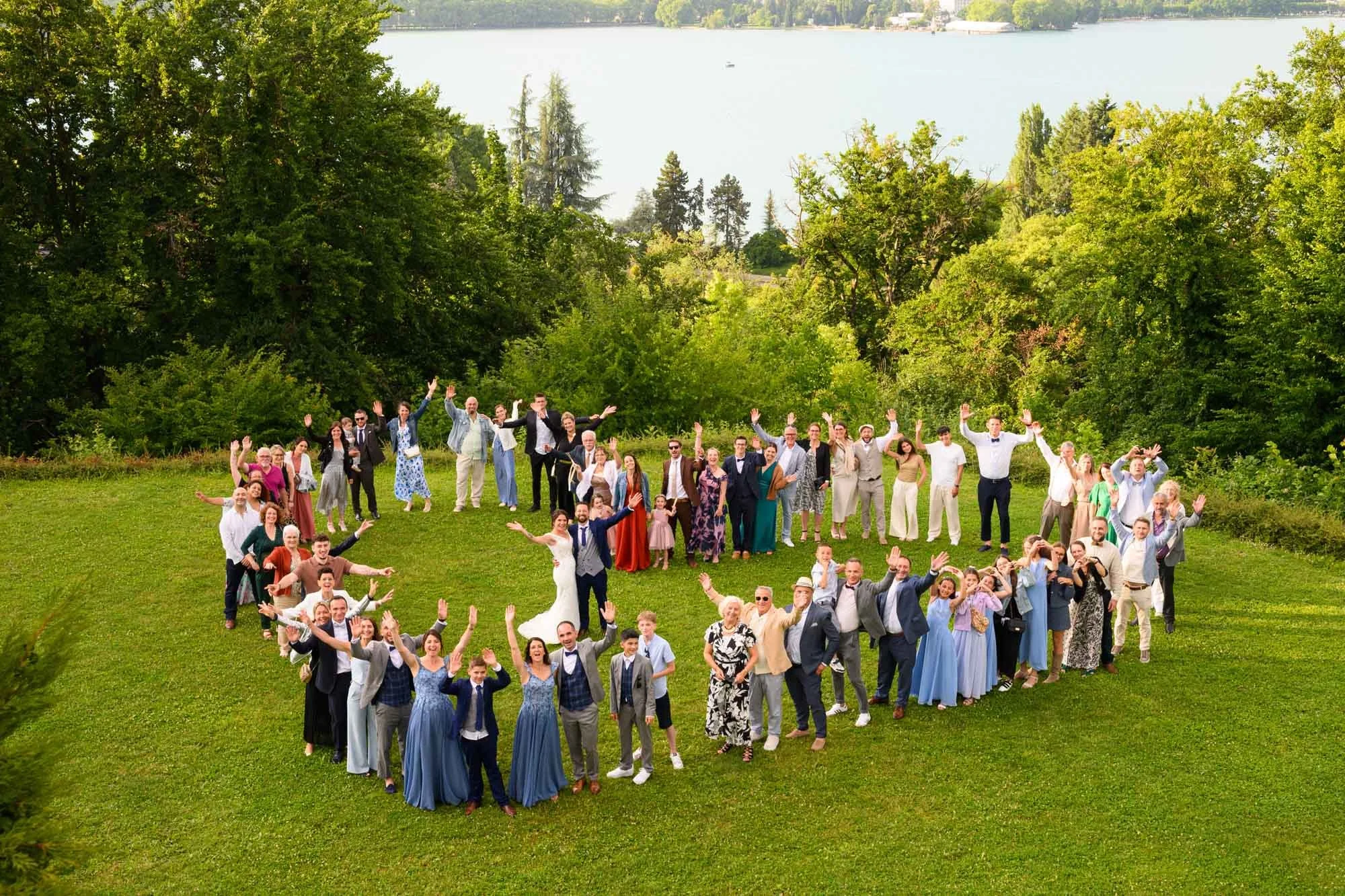Groupe de personnes lors d'un mariage en plein air, formant un cœur sur une pelouse verte, avec des arbres et un lac en arrière-plan.