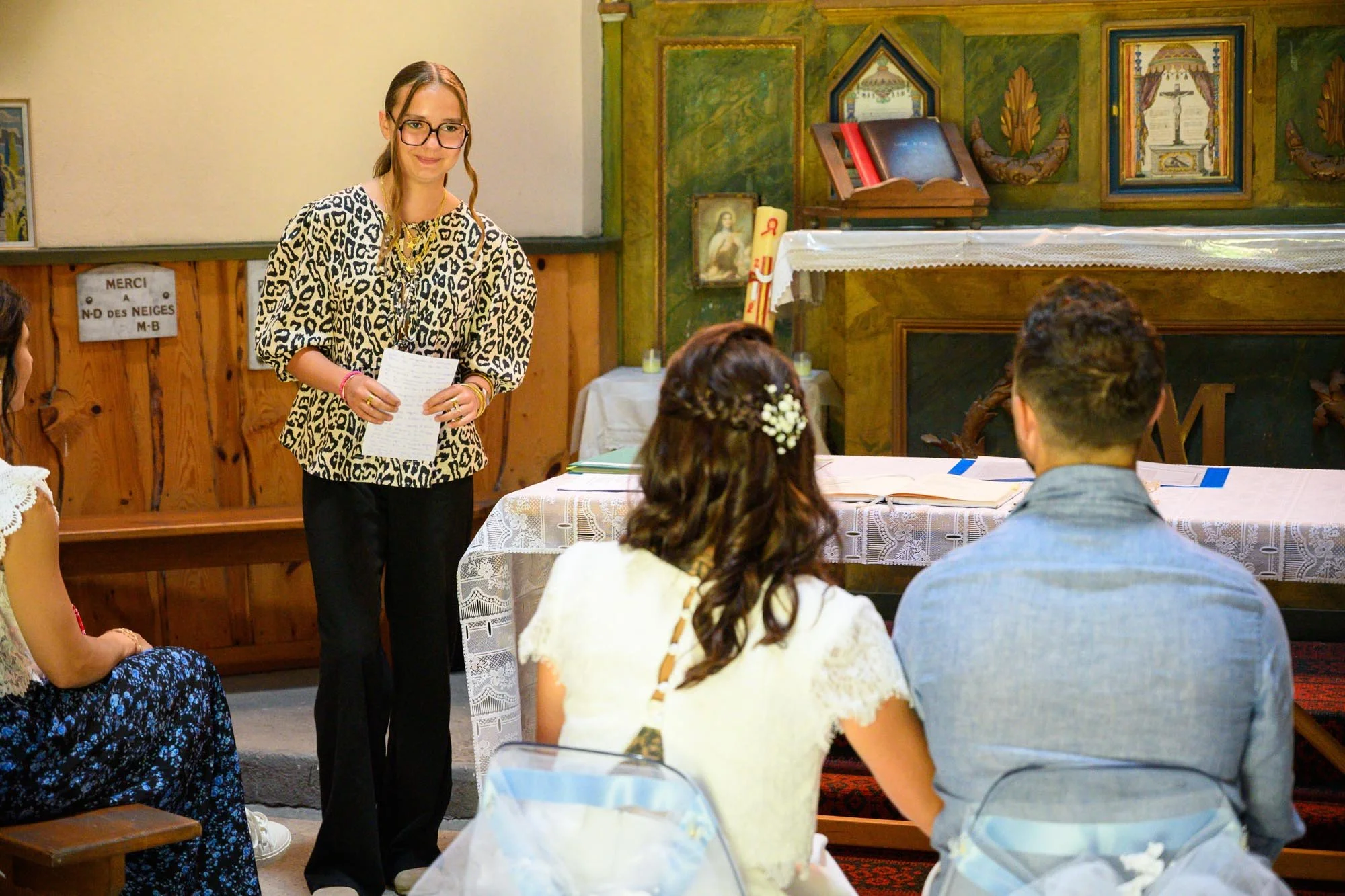 Une jeune femme en lunettes et veste à motif léopard tient un discours dans une église devant plusieurs personnes assises à une table, avec des images religieuses en arrière-plan.