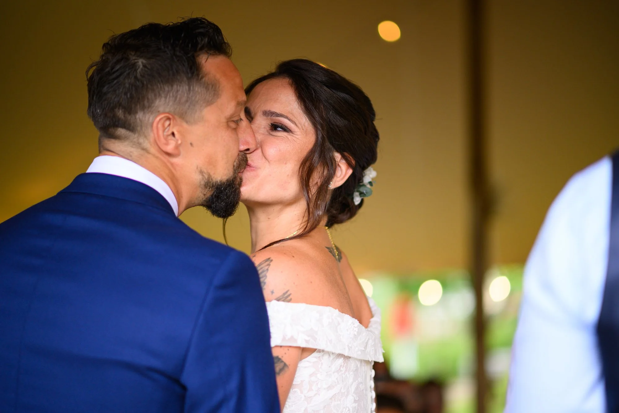 Un couple s'embrasse lors d'une cérémonie de mariage, la femme porte une robe blanche et a des fleurs dans les cheveux, l'homme porte un costume bleu.