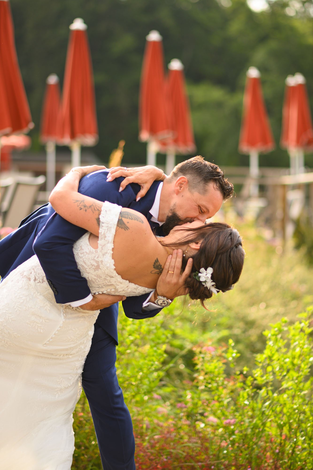 Un couple de mariés en plein baiser lors de leur mariage en extérieur, avec des parasols rouges en arrière-plan.