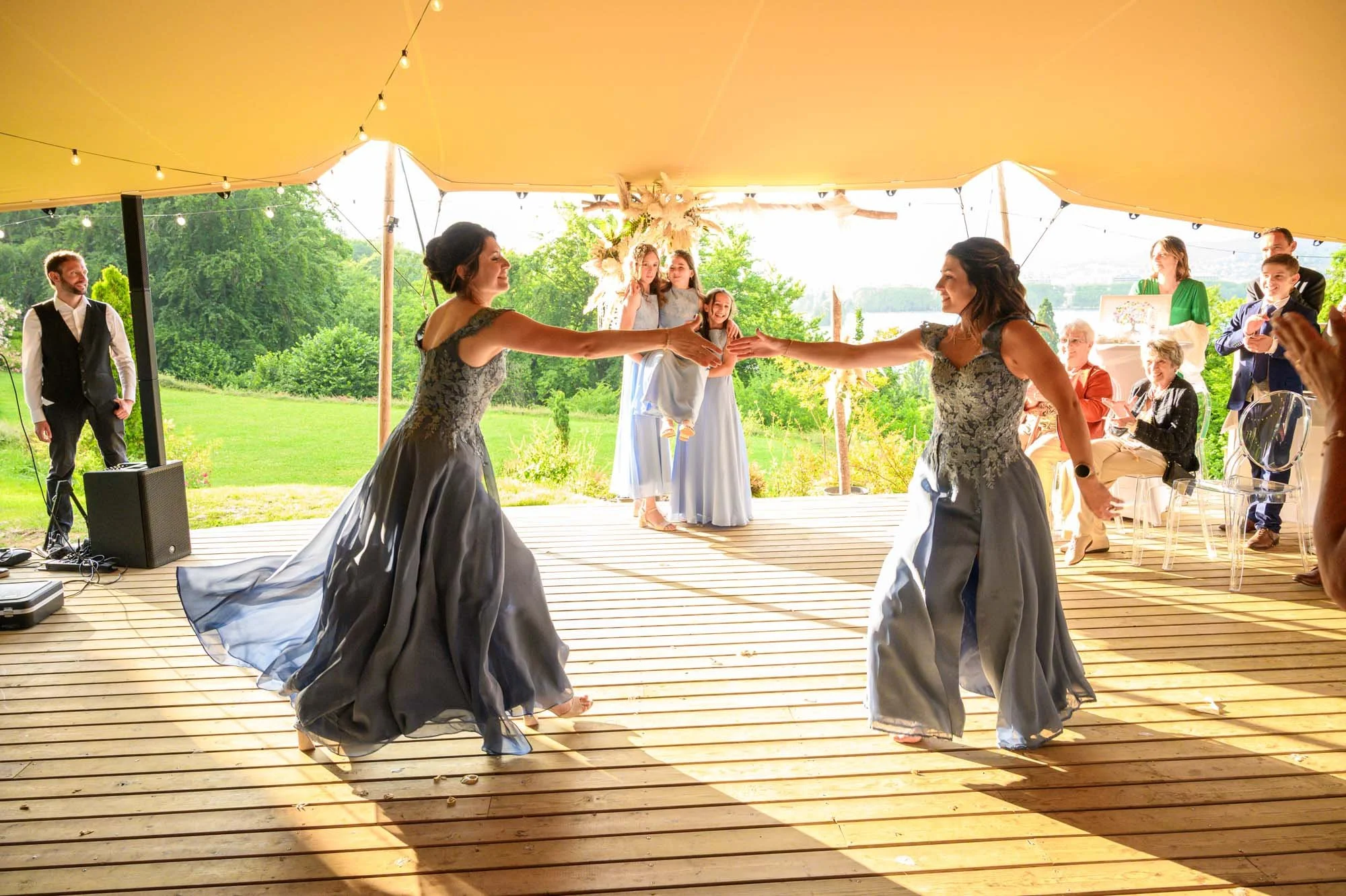 Deux femmes dansant lors d'un mariage en extérieur sous une tente jaune, avec des invités assis en arrière-plan.