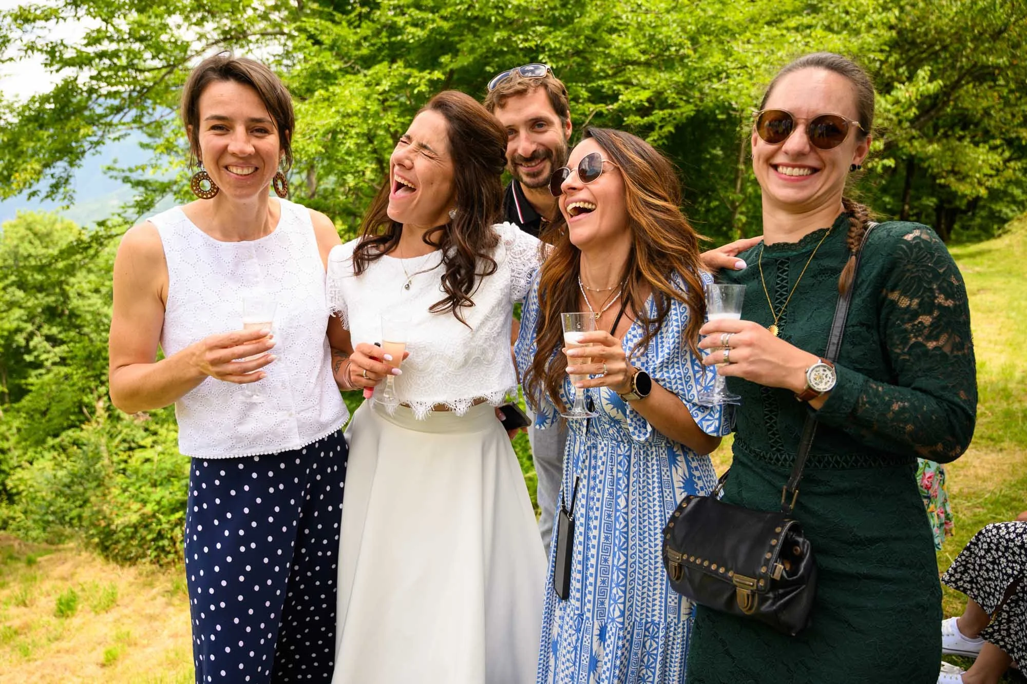 Groupe de six personnes souriantes, tenant des verres, lors d'une fête en extérieur dans un environnement verdoyant.