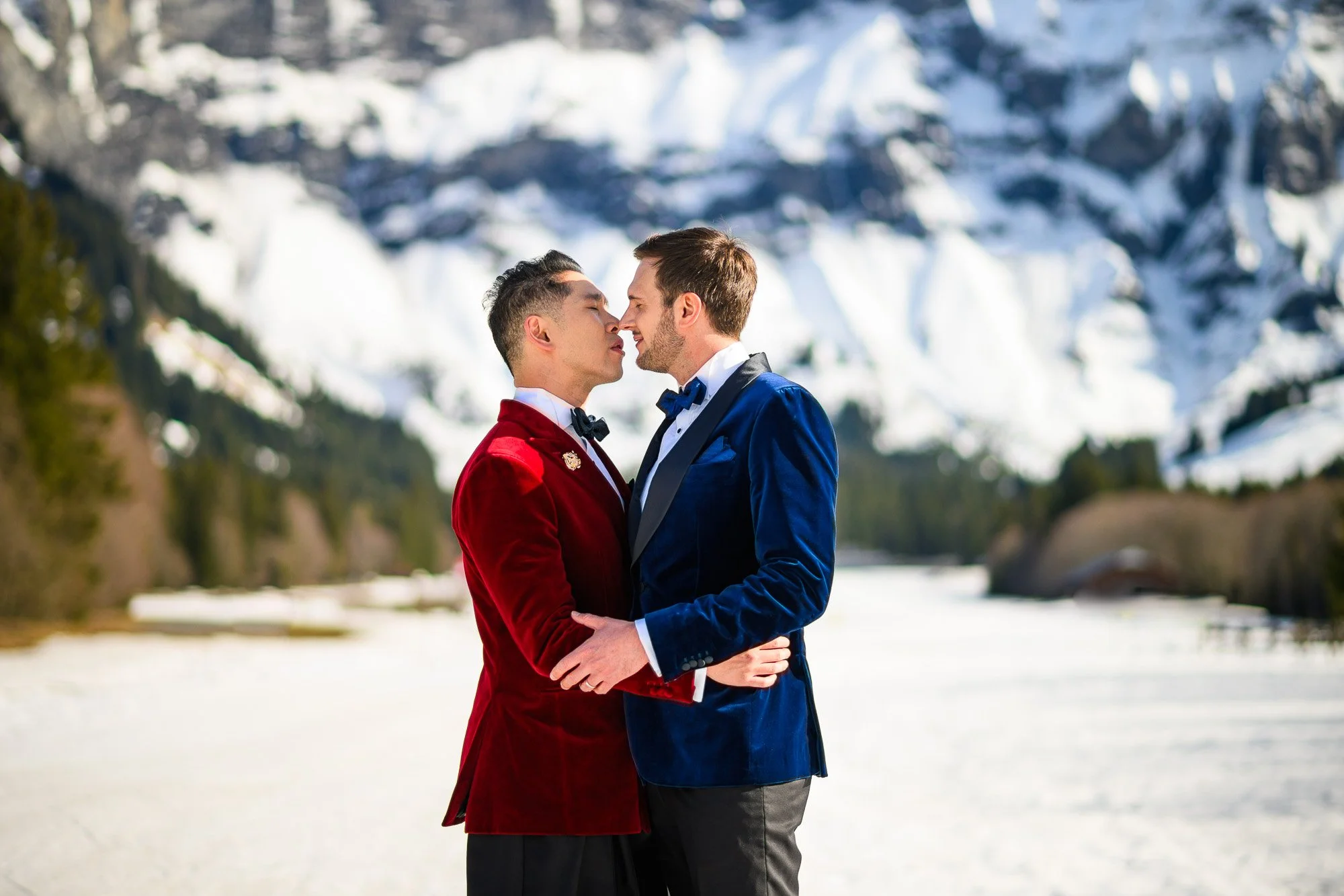 Deux hommes habillés en smokings se tiennent très proches, sur un fond de paysage enneigé avec des montagnes et des arbres, dans une scène romantique.