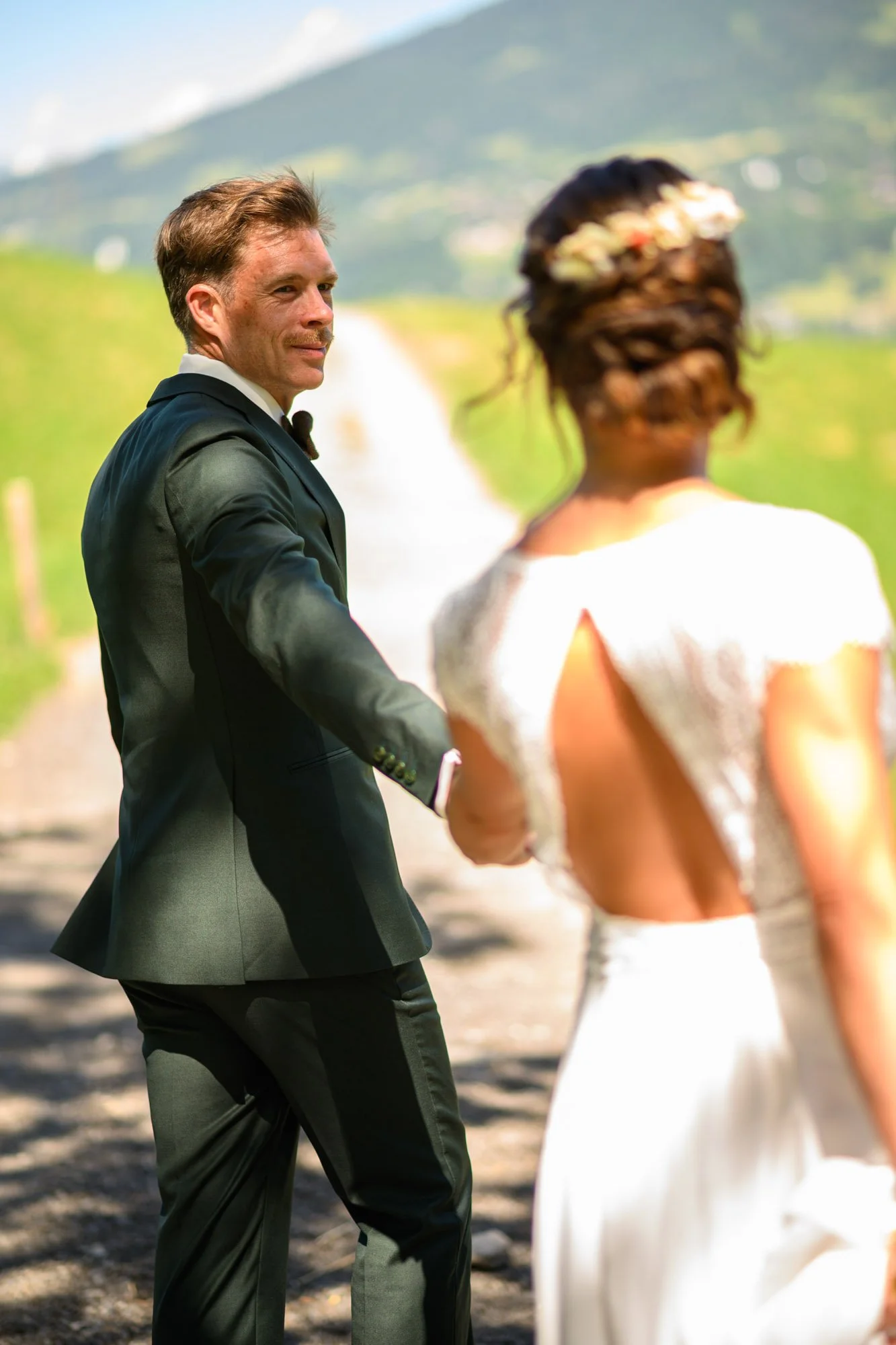 Un couple de mariage en plein air, l'homme en costume noir et la femme en robe de mariage blanche, dans un paysage verdoyant avec des montagnes en arrière-plan.