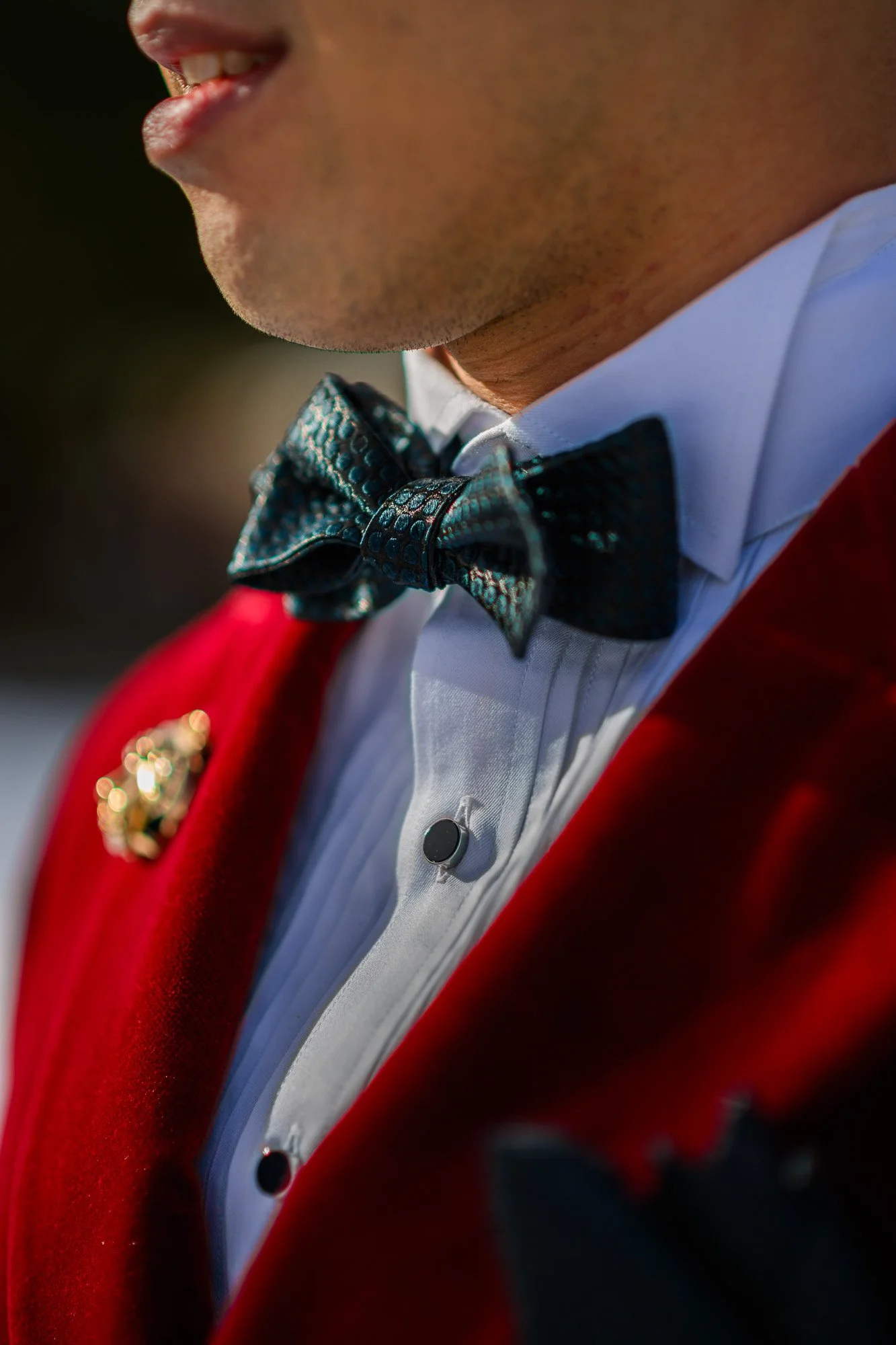 Gros plan sur un homme portant un nœud papillon bleu, une chemise blanche avec boutons noirs, une veste rouge et un bouton de décor en or.