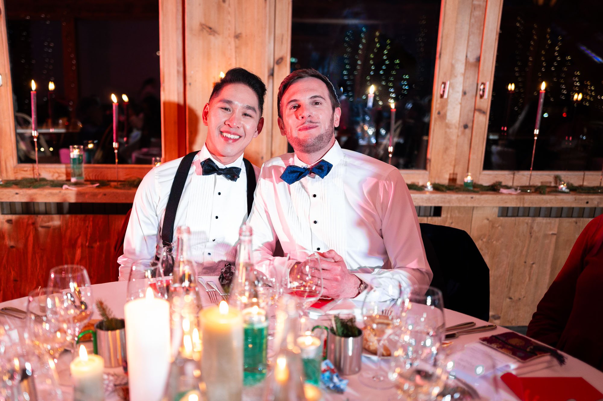 Deux hommes habillés en smokings avec des nœuds papillon assis à une table de dîner de fête, avec bougies et décorations festives, dans une ambiance chaleureuse et festive.