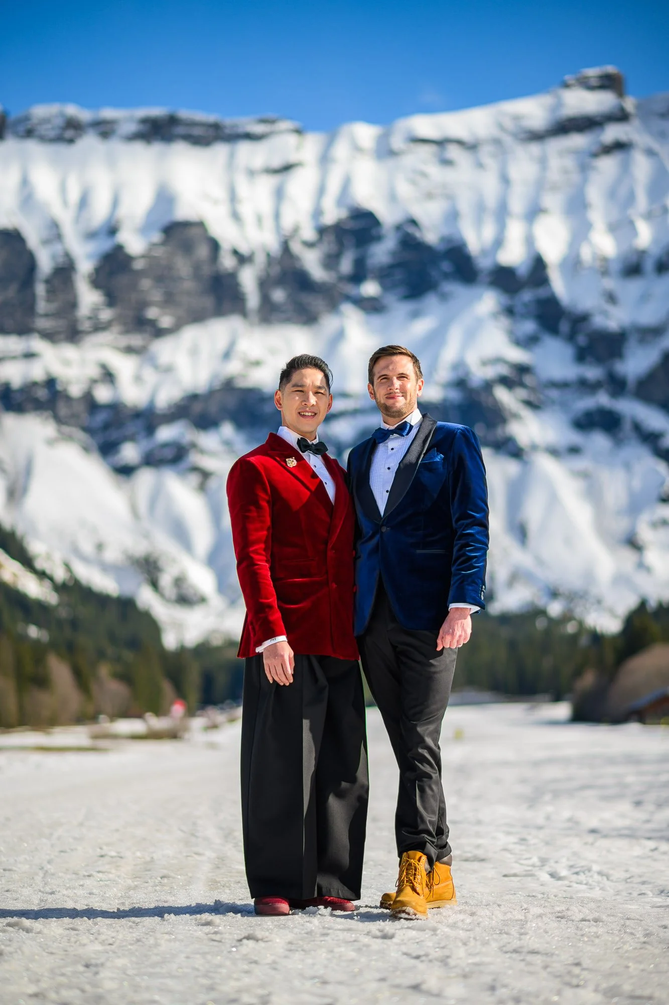 Deux hommes habillés en smokings colorés (rouge et bleu) se tiennent côte à côte dans un paysage enneigé avec des montagnes enneigées en arrière-plan, sous un ciel bleu.