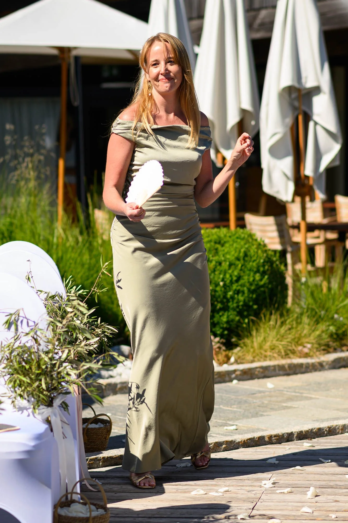 Une femme portant une robe longue de couleur beige, souriant, tenant une plume blanche dans sa main, en plein air lors d'une cérémonie ou d'un événement.