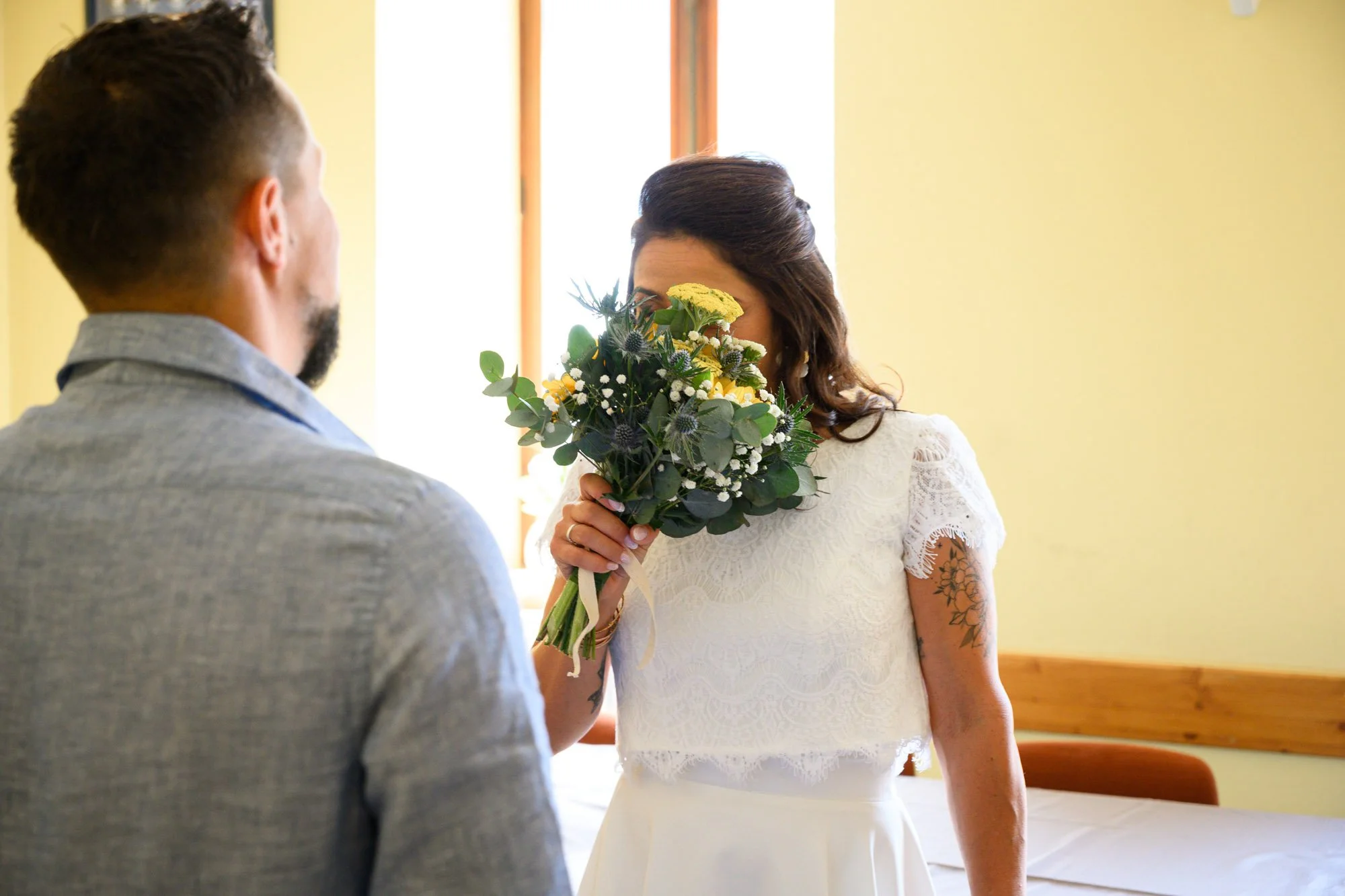 Une femme en robe blanche tenant un bouquet de fleurs devant son visage, lors d'une cérémonie ou d'un moment spécial, avec un homme dont le dos est visible dans une pièce lumineuse.