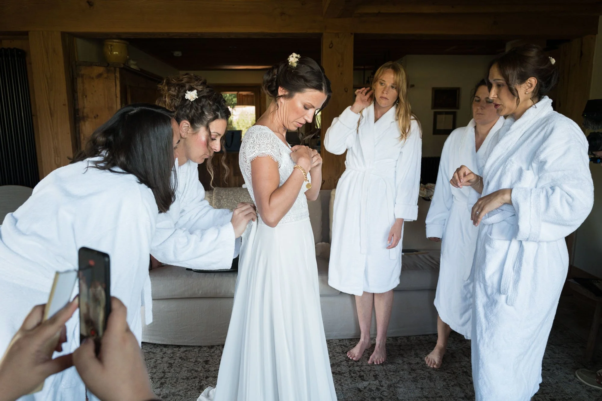 Une mariée en robe de mariage blanche entourée de femmes en peignoirs blancs, probablement ses demoiselles d'honneur, dans une pièce en bois, en préparation pour un mariage.
