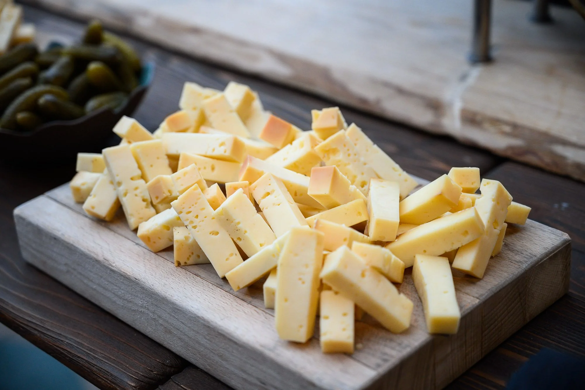 Fromage en morceaux sur une planche en bois, accompagné de cornichons dans un bol noir à côté.