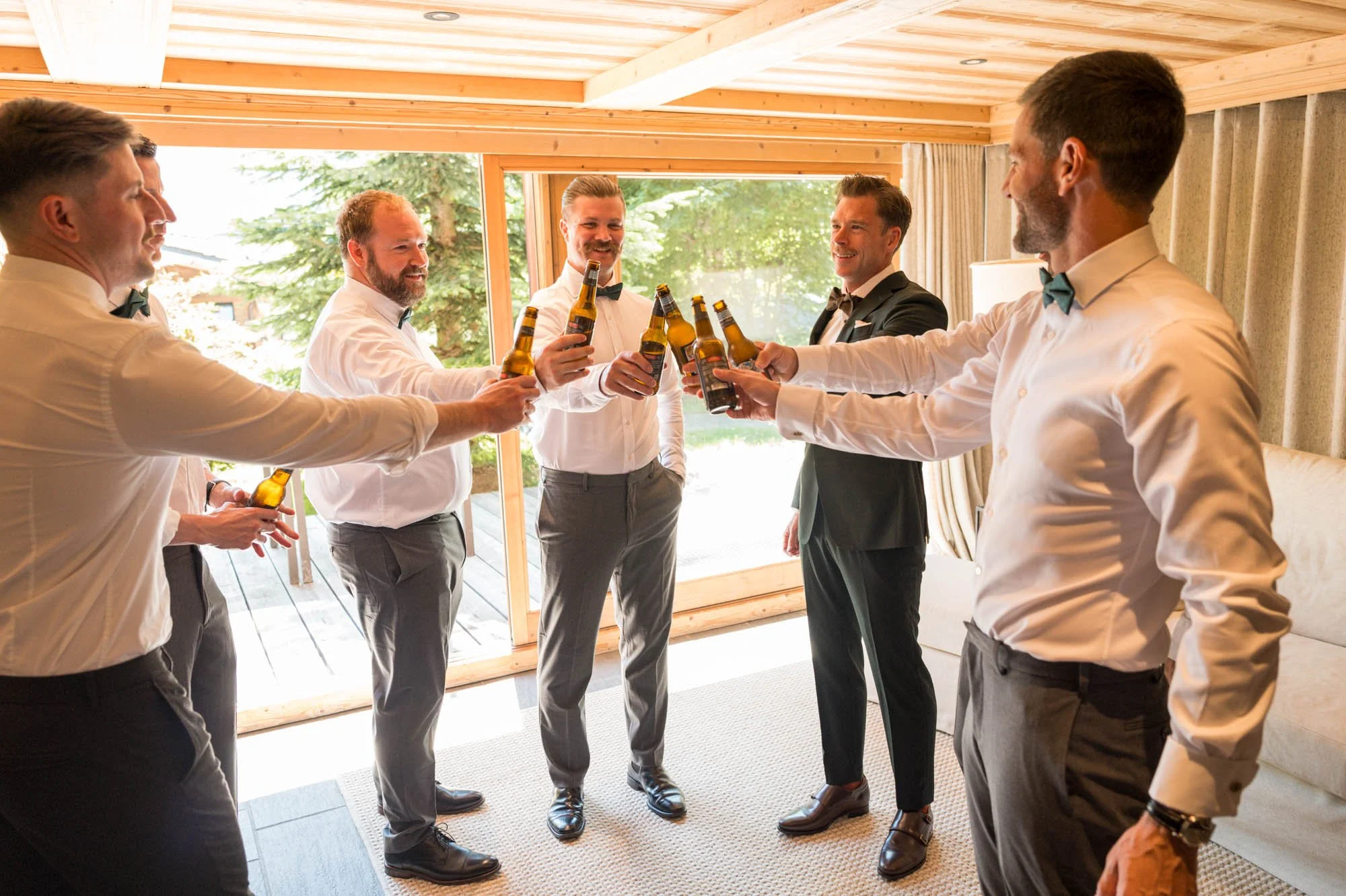 Groupe d'hommes habillés en costumes formels autour d'une table, levant des bouteilles de bière en célébration dans une pièce lumineuse avec vue sur la nature extérieure.