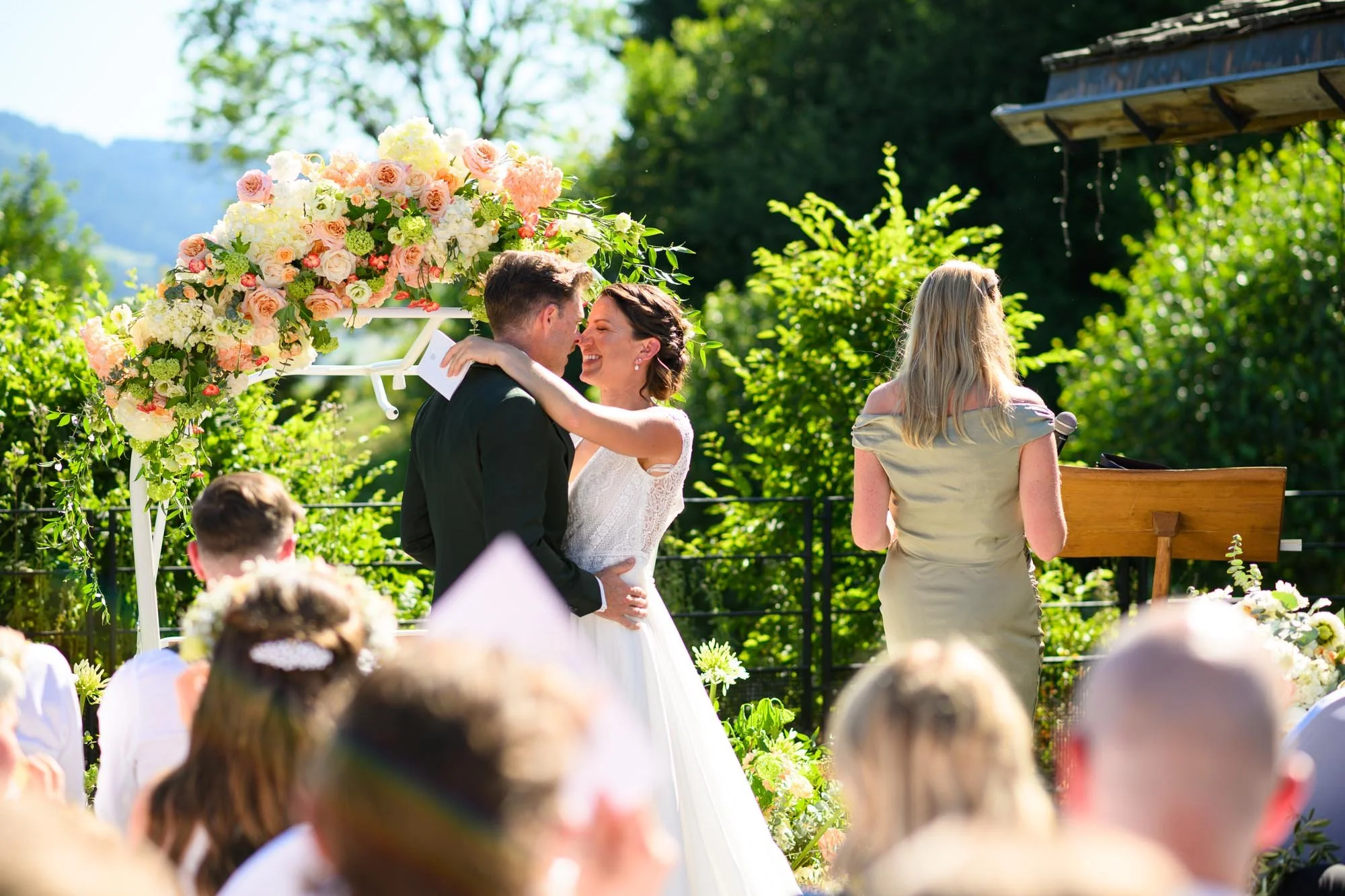 Un couple de mariés s'embrassent sous un arc de fleurs lors d'une cérémonie en extérieur, entourés d'invités et d'une officiante.