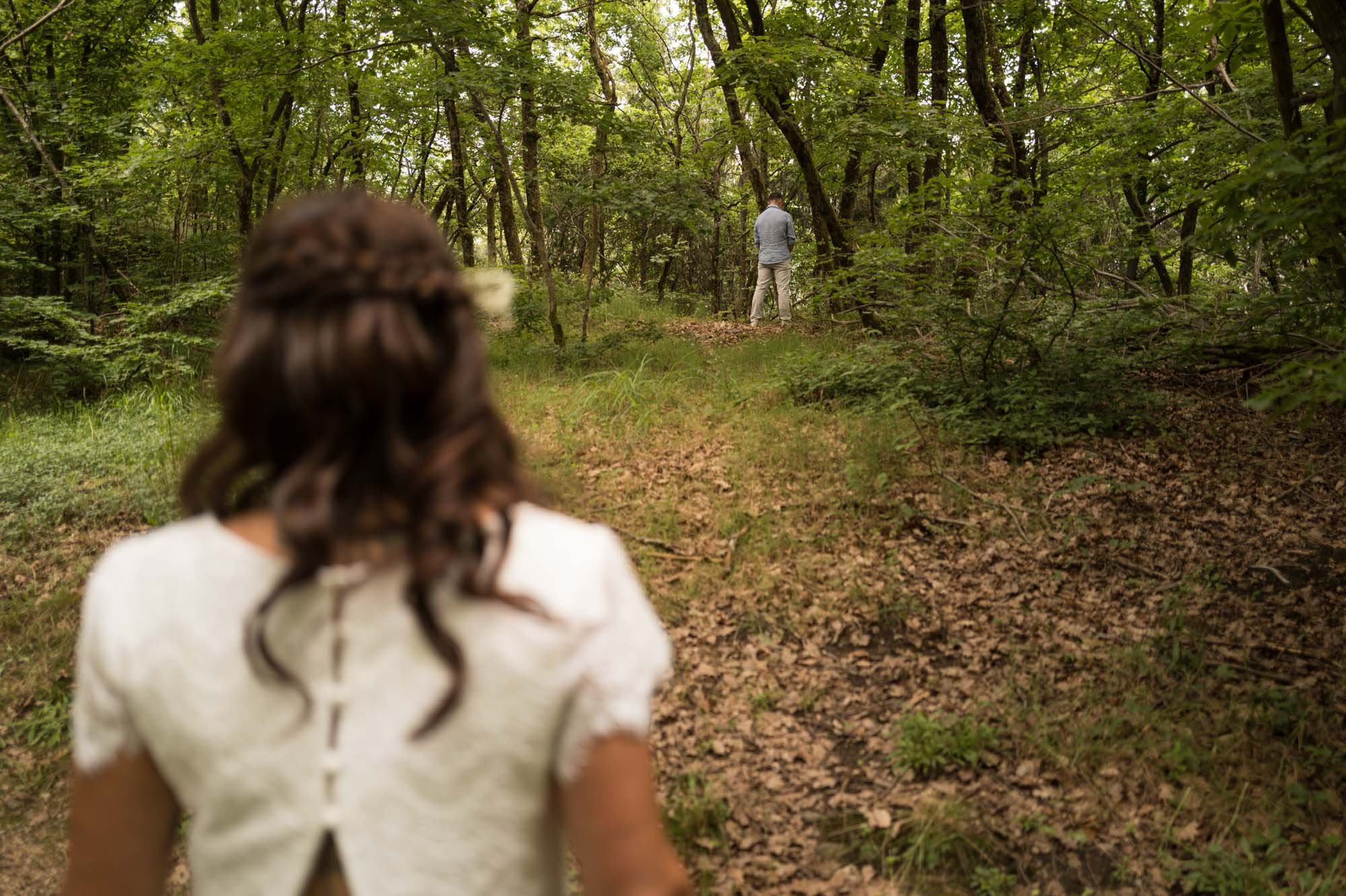 Une femme avec des cheveux bouclés portant une robe blanche observe un homme dans un bois verdoyant. L'homme est agenouillé et semble être en train de faire une proposition ou d'attendre une réponse.