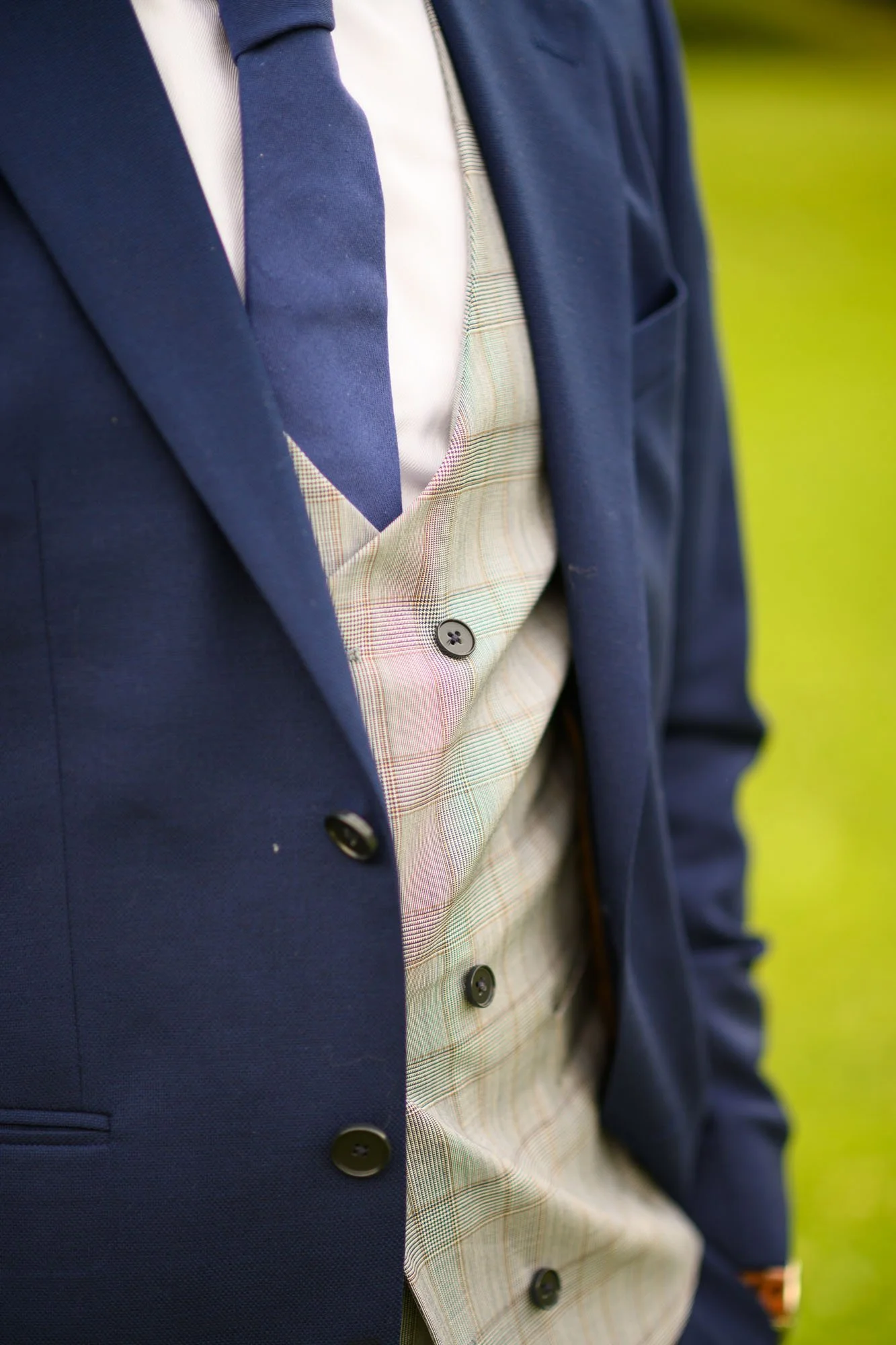Un homme porte un costume bleu foncé avec une veste et un pantalon, une chemise blanche, une cravate bleue et un gilet à carreaux beige avec des boutons noirs.