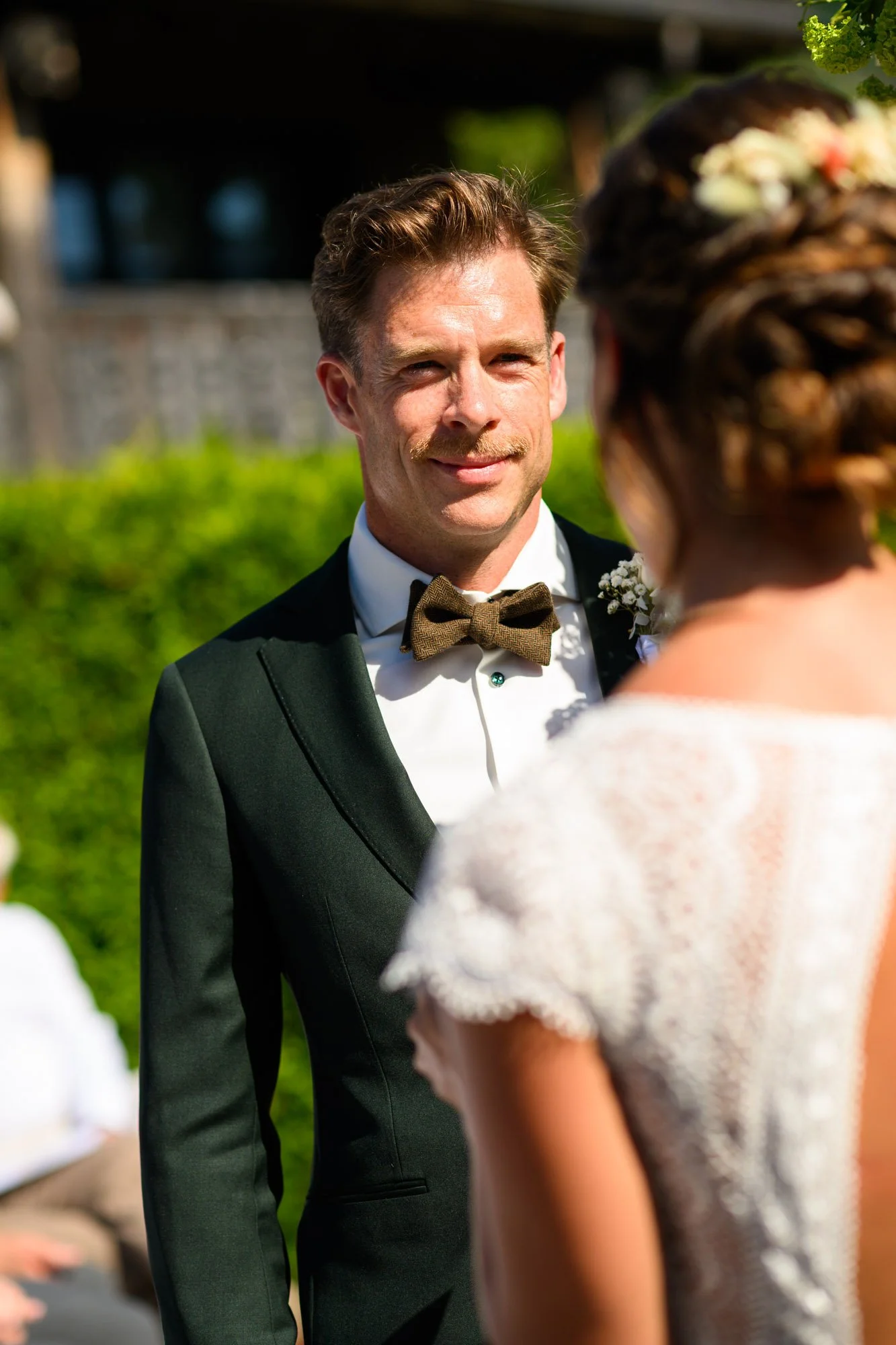 Un homme en costume de mariage regardant une femme lors d'une cérémonie en plein air, avec un fond verdoyant.