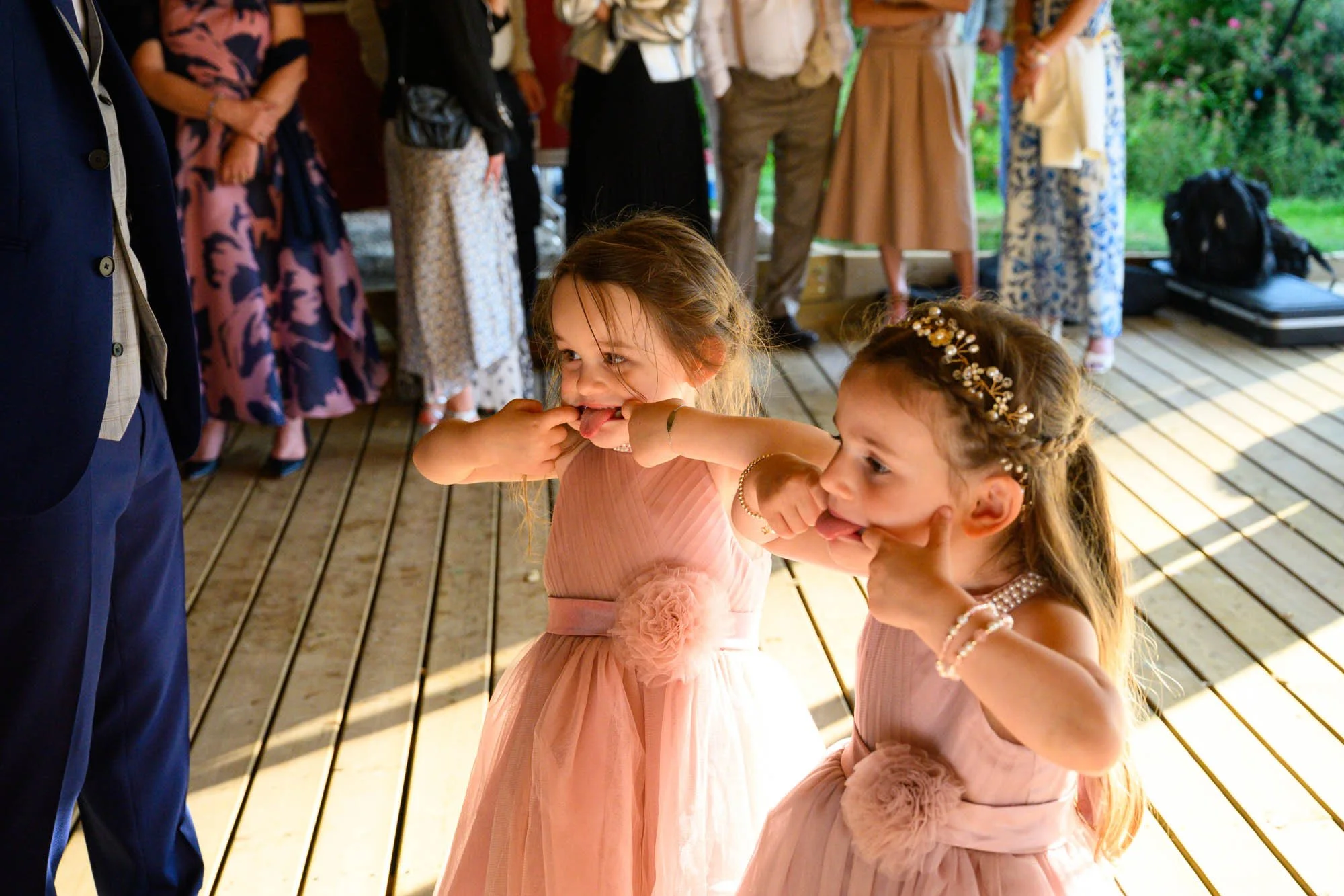Deux jeunes filles en robes roses se taquinant, en pointant leur langue, lors d'une fête ou d'un mariage, avec des invités en arrière-plan.