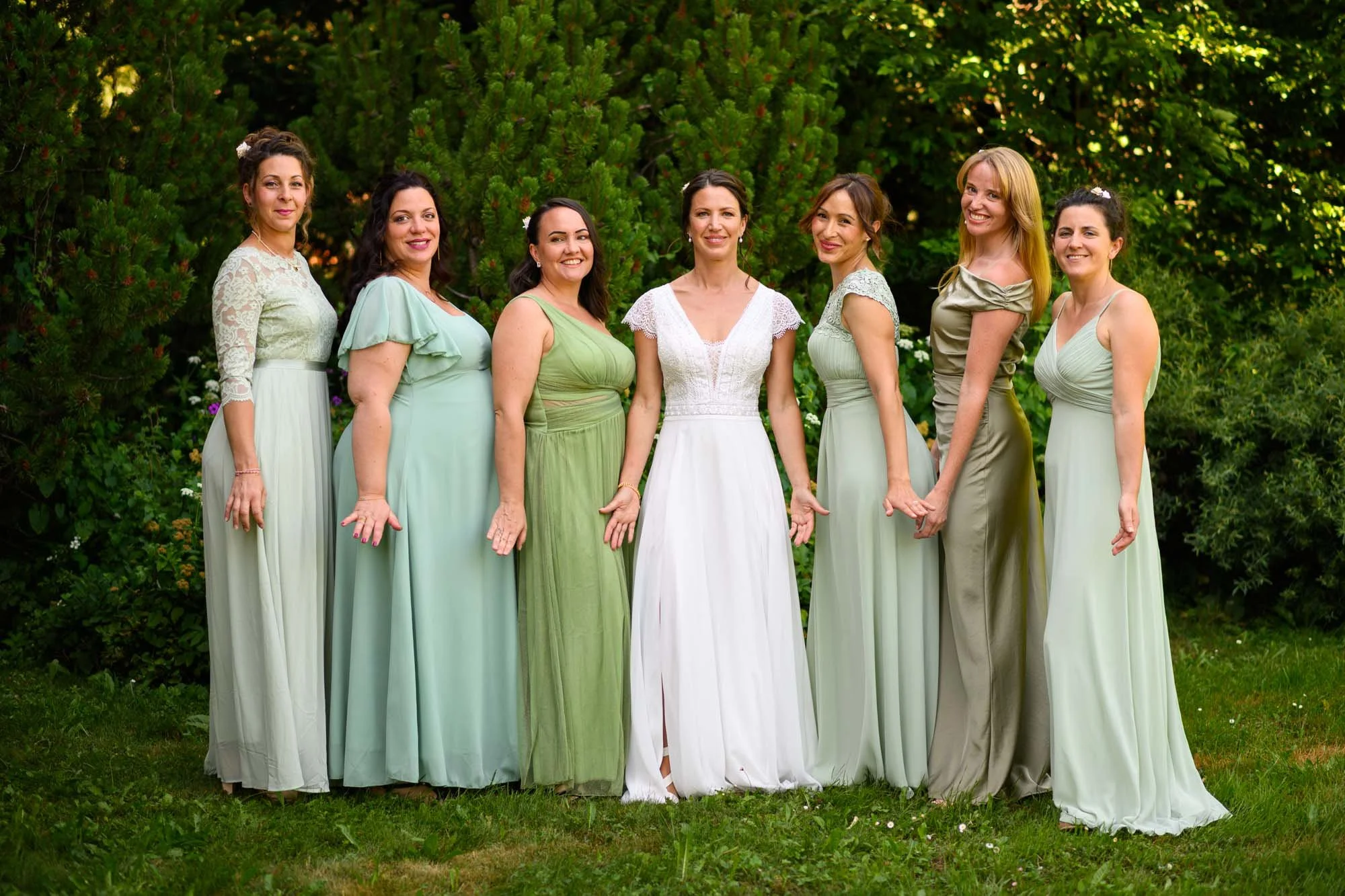 Groupe de sept femmes en robes de cérémonie, debout sur une pelouse verte avec un arbuste en arrière-plan, lors d'une occasion spéciale comme un mariage.